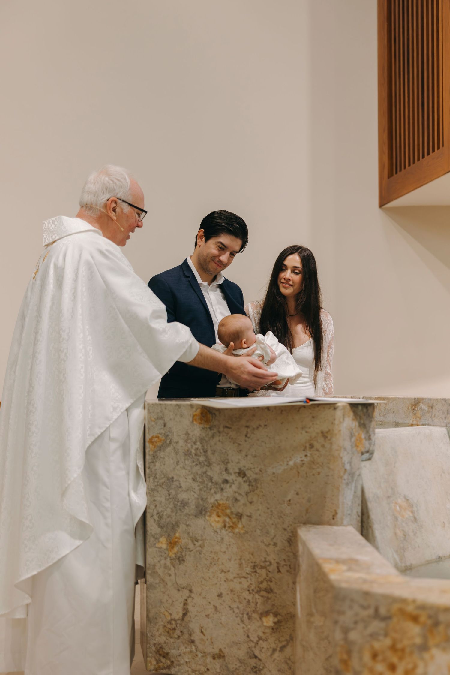 Smiling priest baptizes infant at marble font as proud parents look on in church.