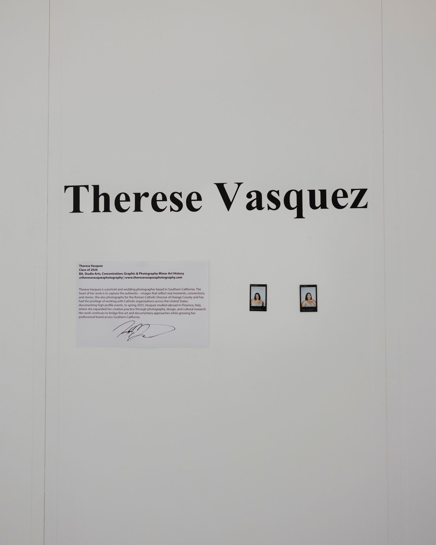 White gallery wall with large text displaying 'Therese Vasquez' and small framed items.