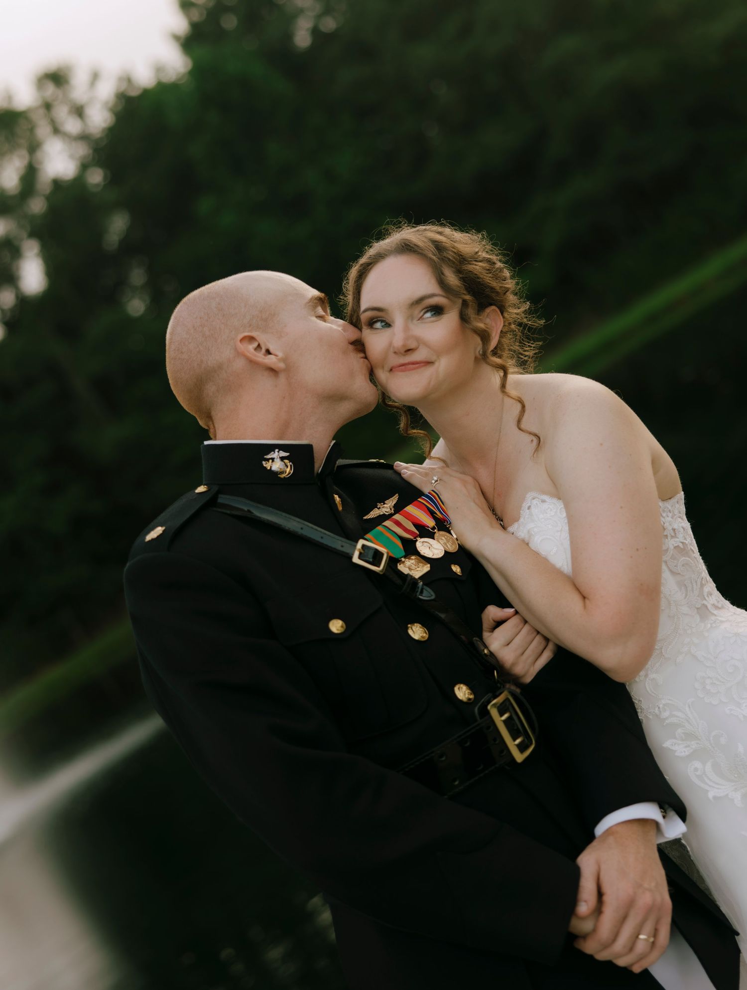 A US Marine in dress uniform kisses his bride on the cheek during their outdoor wedding portrait.