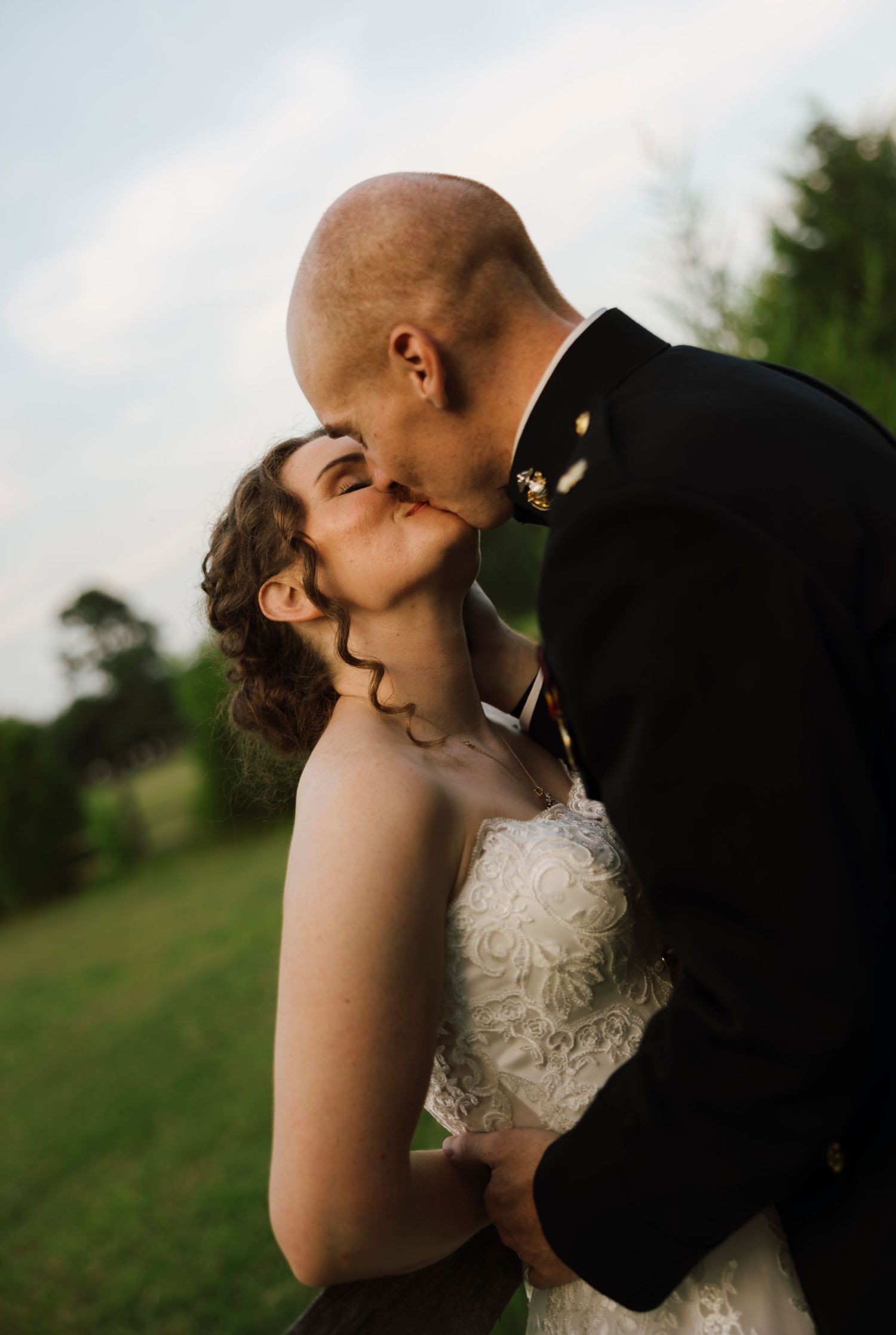 A bride and groom share a romantic kiss outdoors, with the groom wearing a military dress uniform on their wedding day.