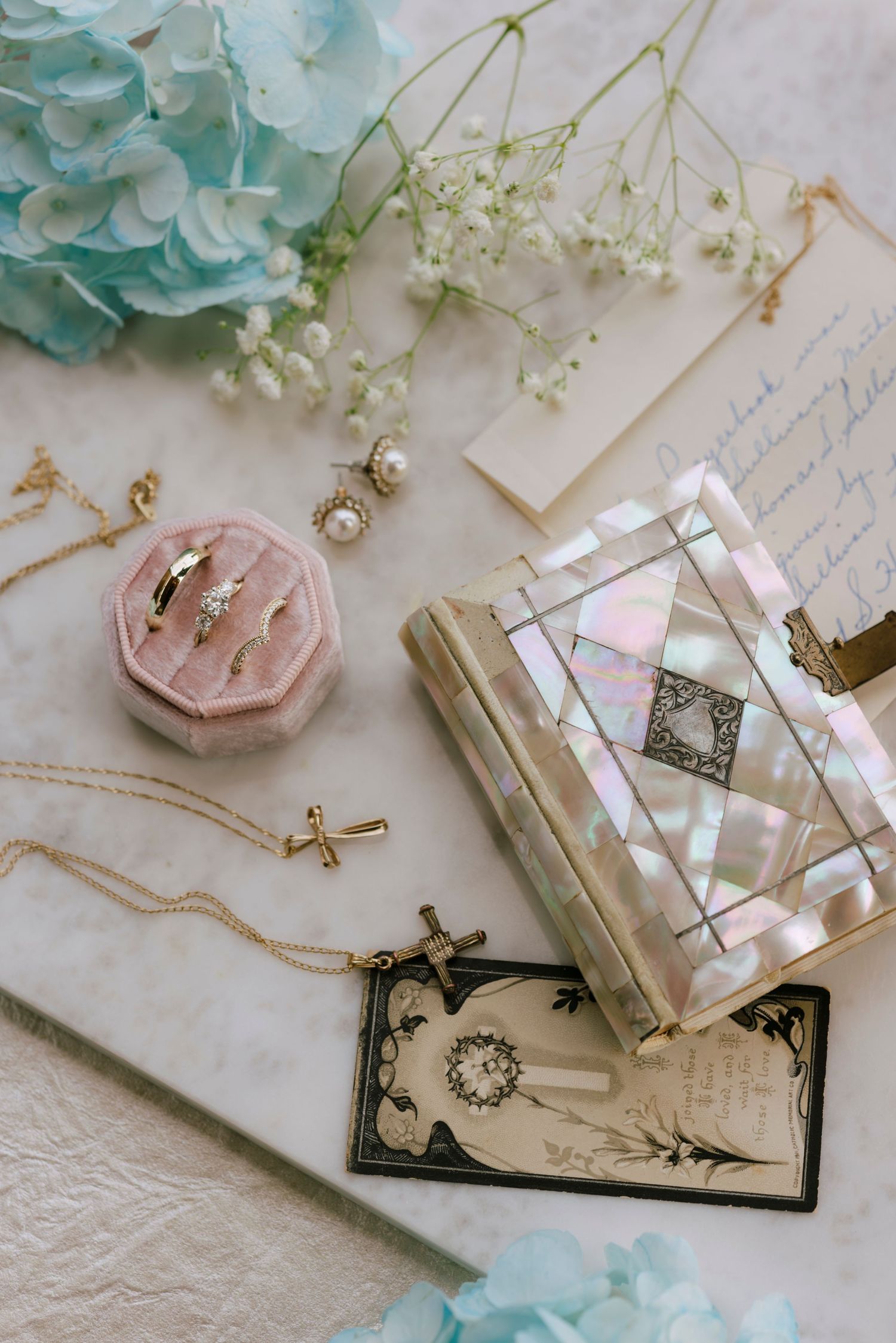 Elegant bridal flat lay with pearl earrings, gold rings in pink velvet box, cross necklace, mother-of-pearl clutch, and hydrangea flowers.