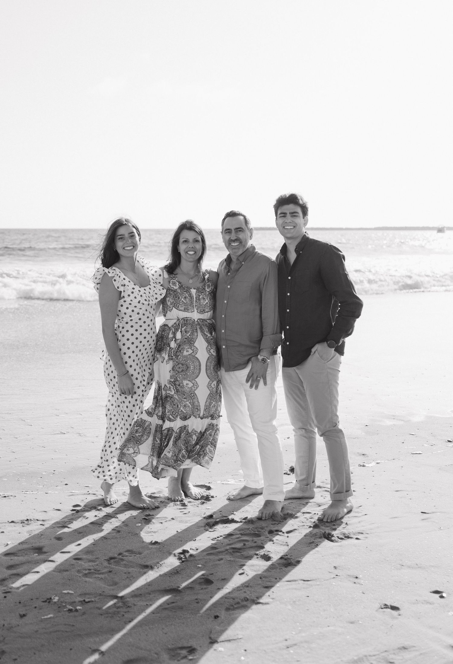 Black and white family portrait of four smiling on a sunny beach with ocean waves.