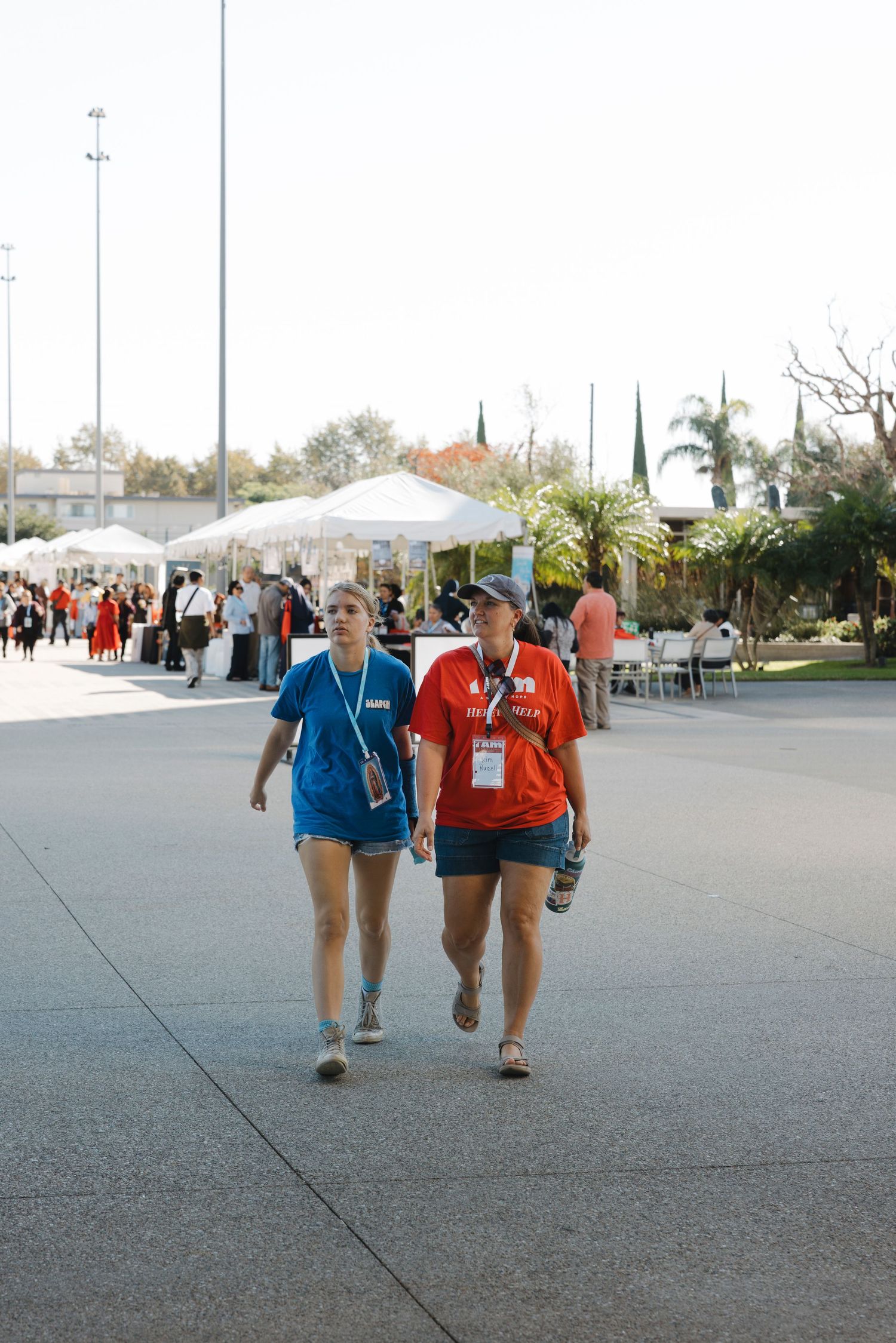 People walking under white tents at an outdoor event with palm trees in the background.