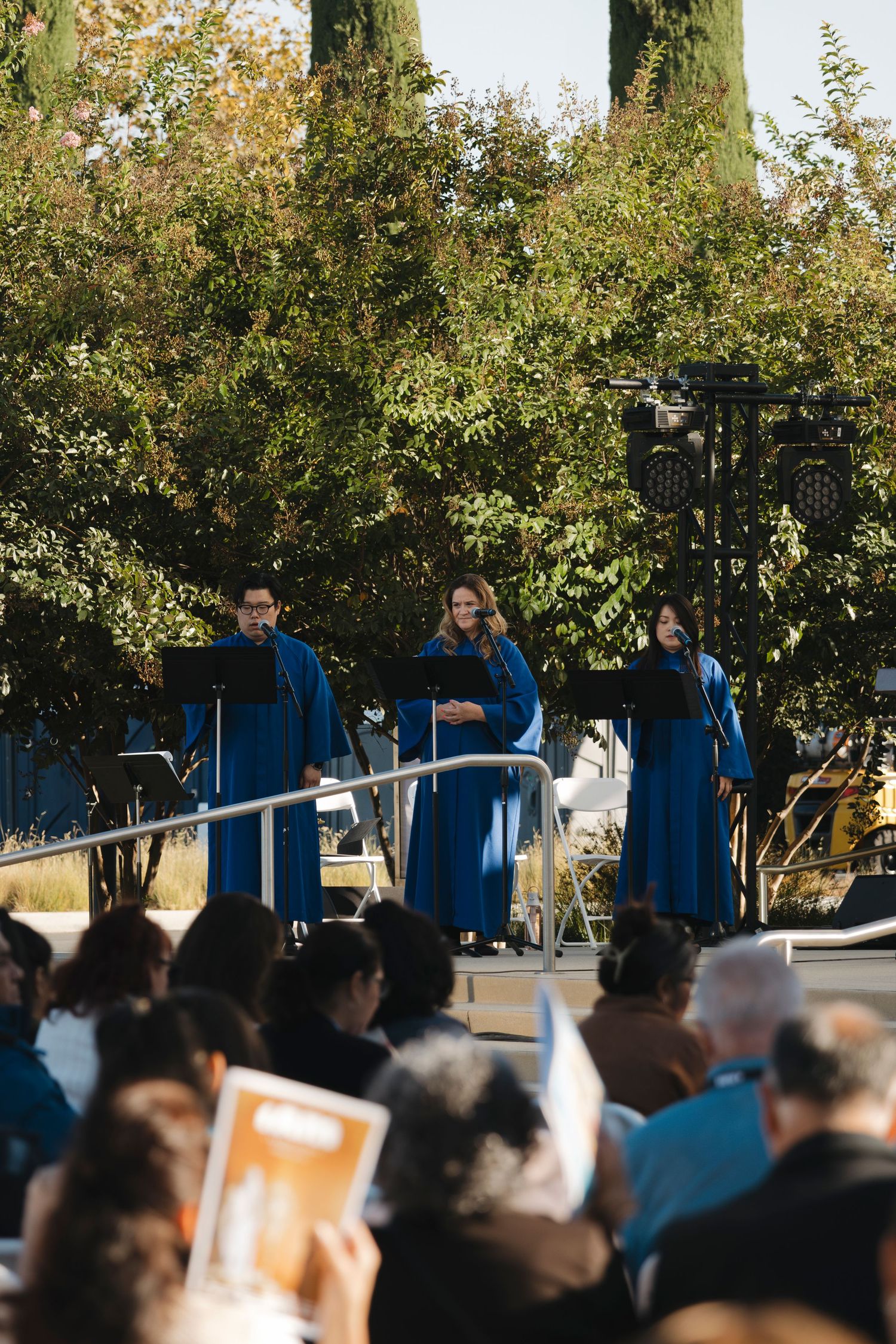 Outdoor gathering with crowd seated beneath trees and blue-uniformed attendants.