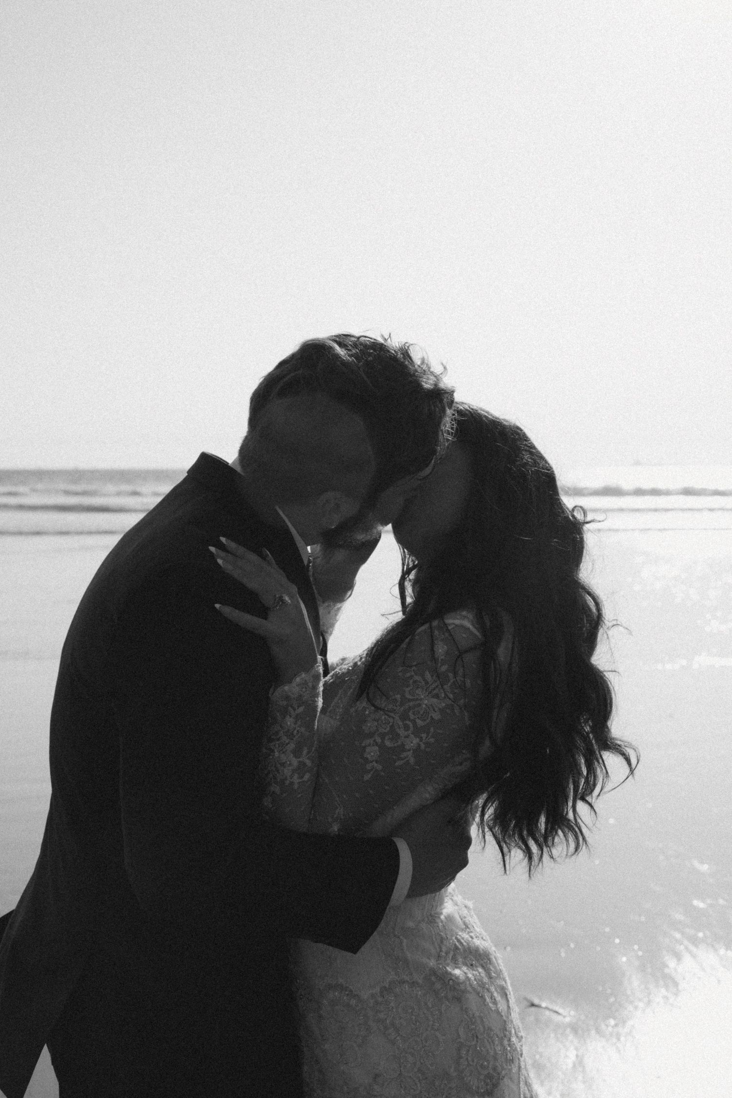 Black and white silhouette of newlyweds embracing on beach with ocean backdrop.