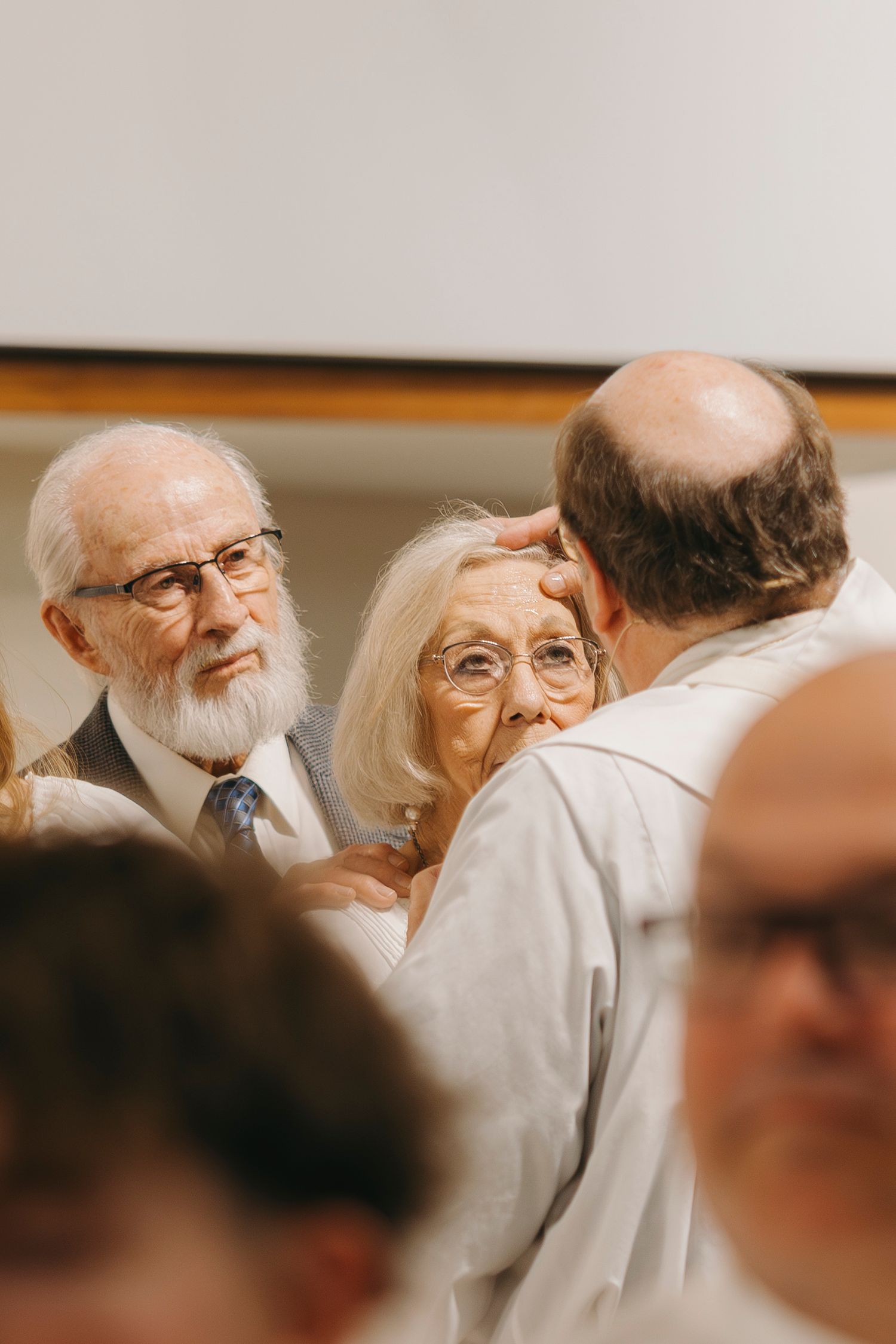 Elderly couple with glasses receiving a blessing from a priest during a religious ceremony indoors.