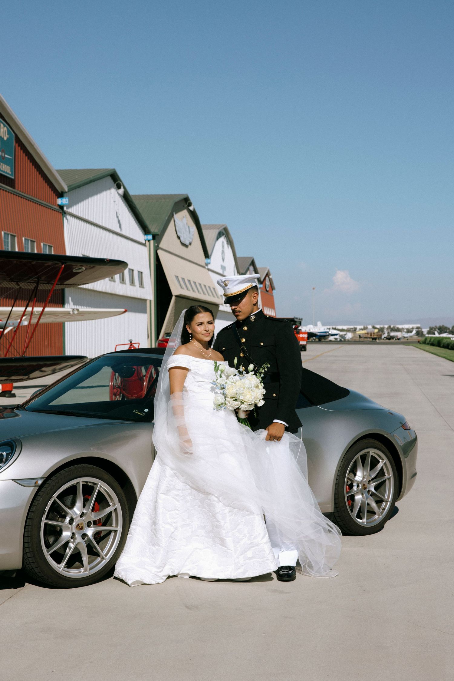 Wedding couple poses with luxury sports cars outside modern buildings.