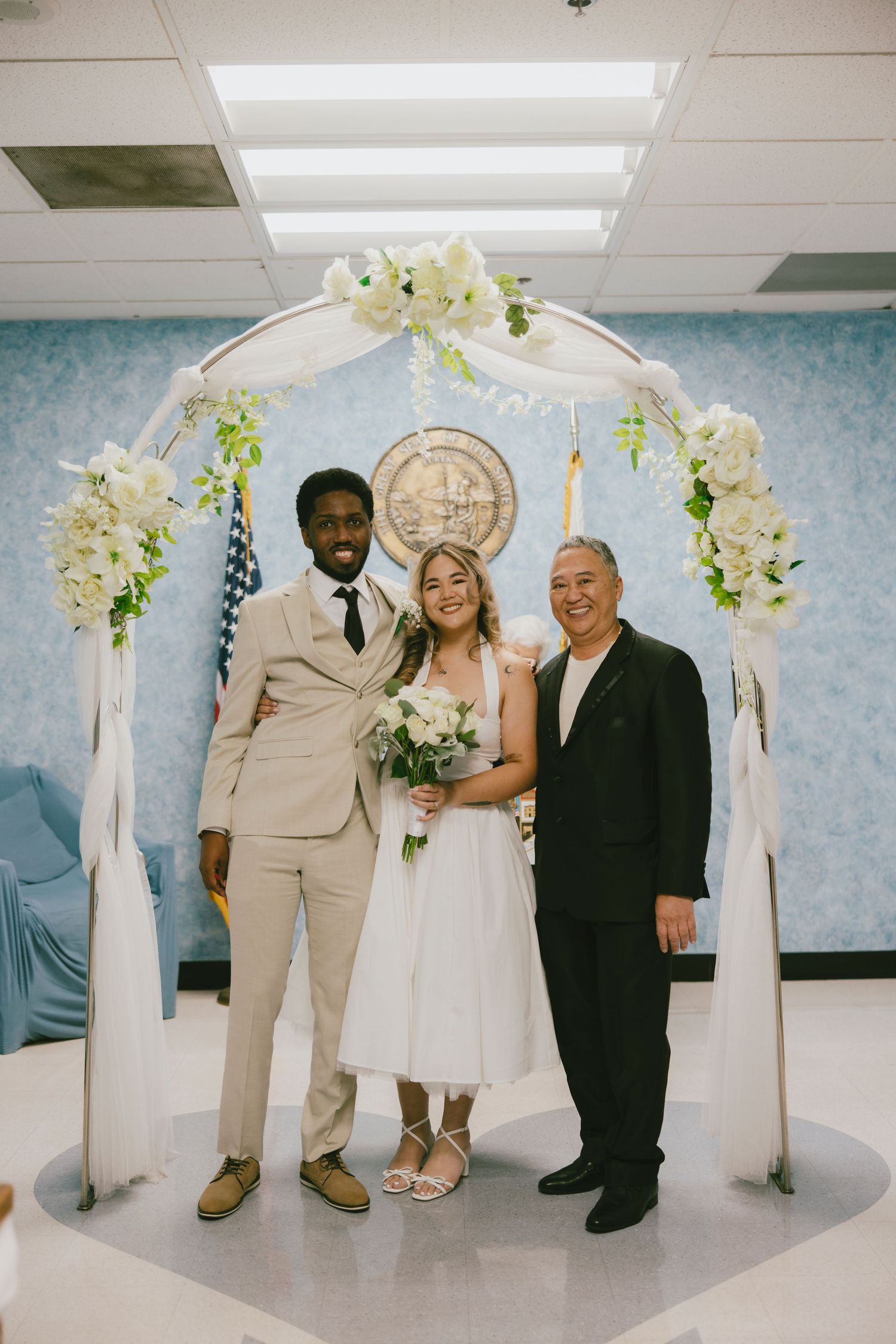 Wedding couple poses with an officiant under a floral arch during their indoor ceremony.