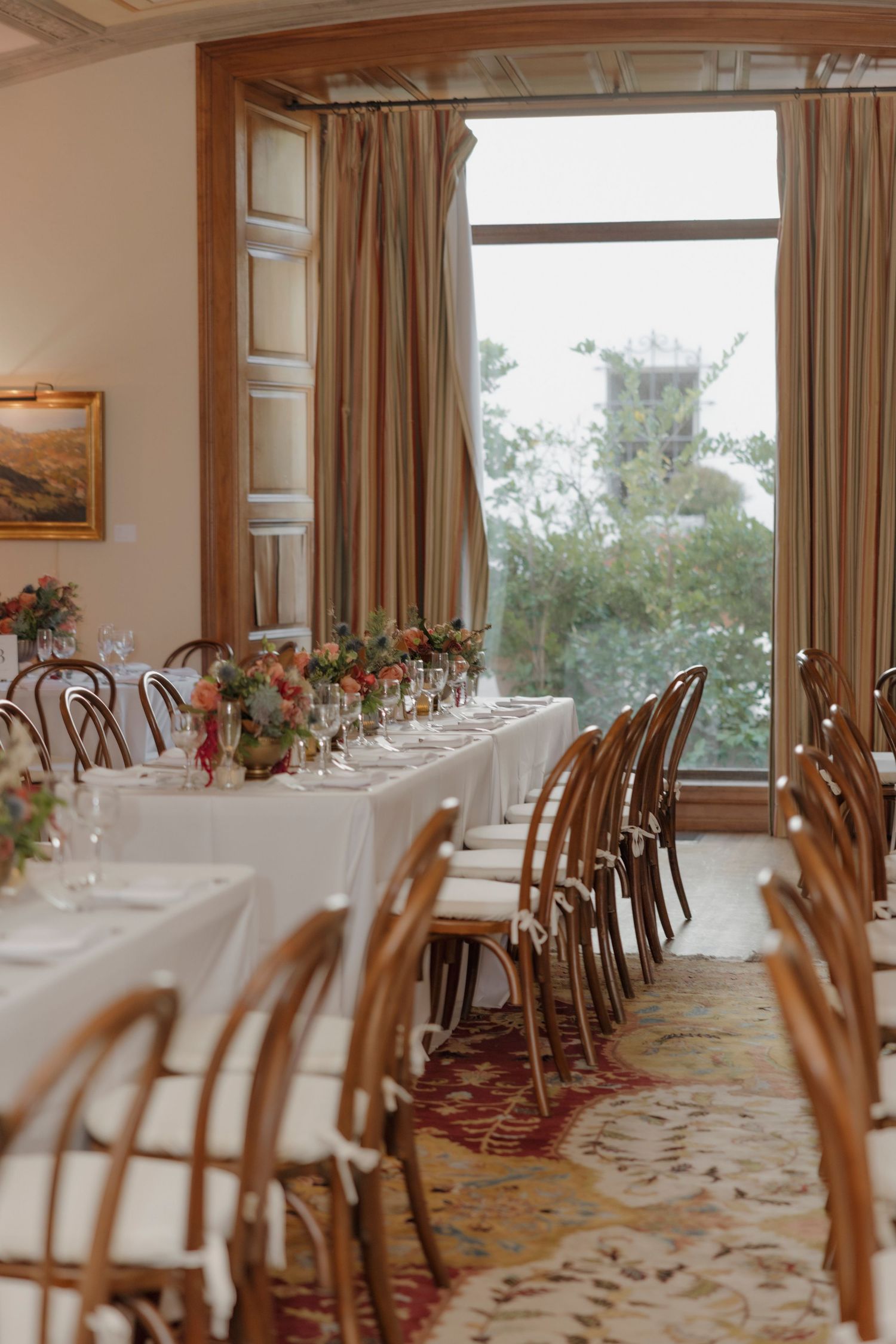 Spacious dining room with wooden chairs and white-clothed tables arranged in rows near large windows.