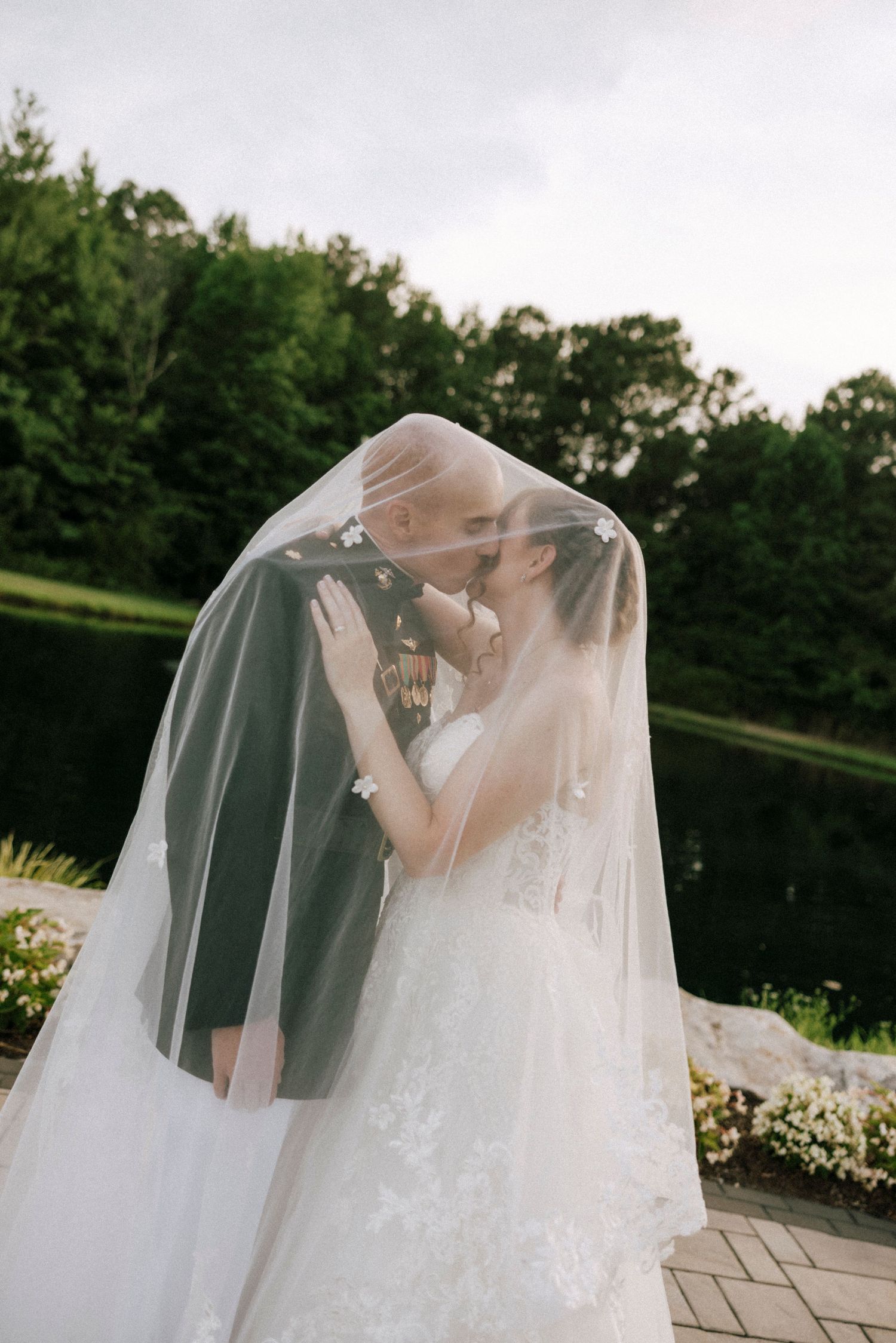Bride and groom sharing a romantic kiss under a flowing white veil in an outdoor garden wedding setting.