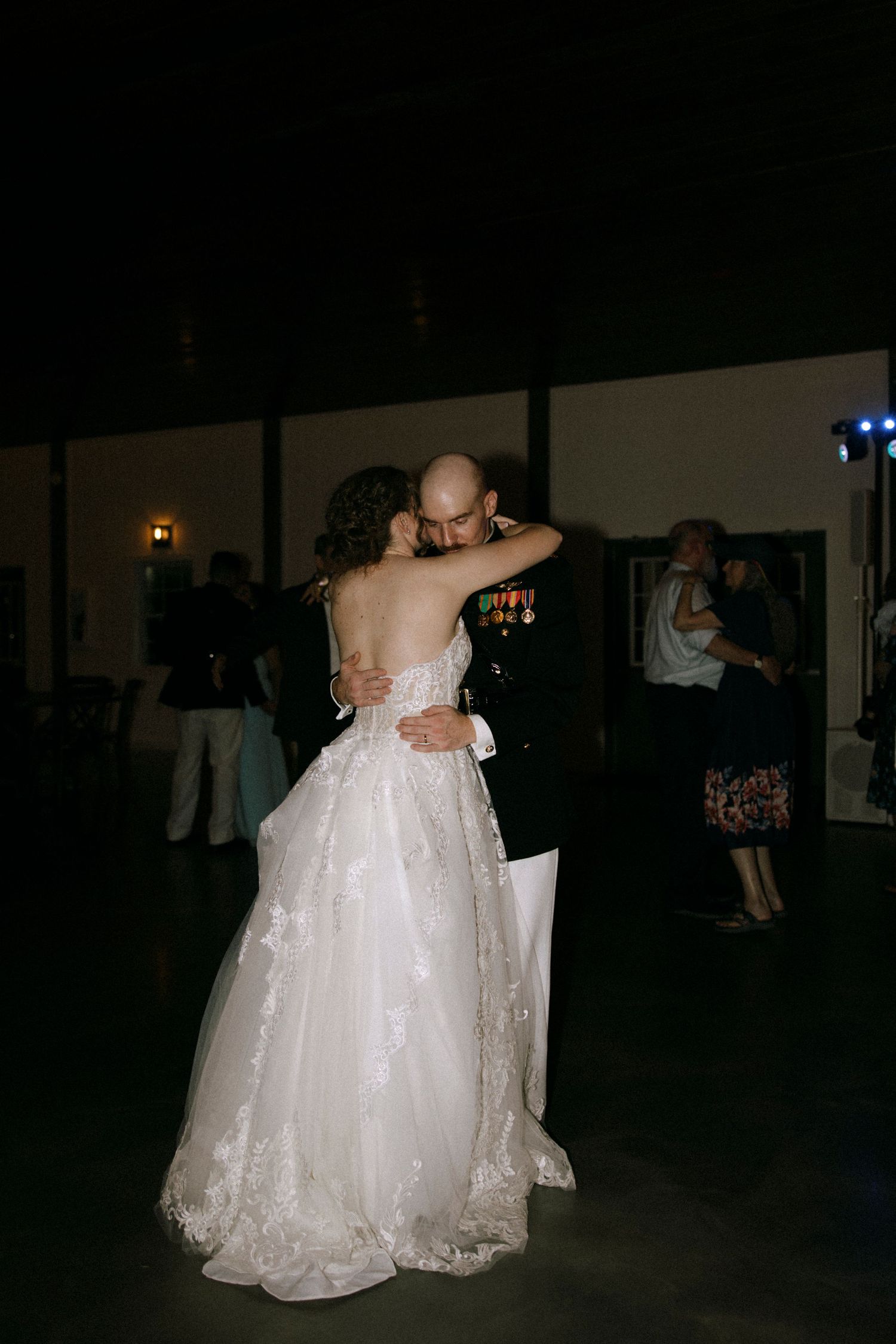 Bride and groom share their first dance, bride in lace gown, groom in military uniform, at a dimly lit wedding reception.