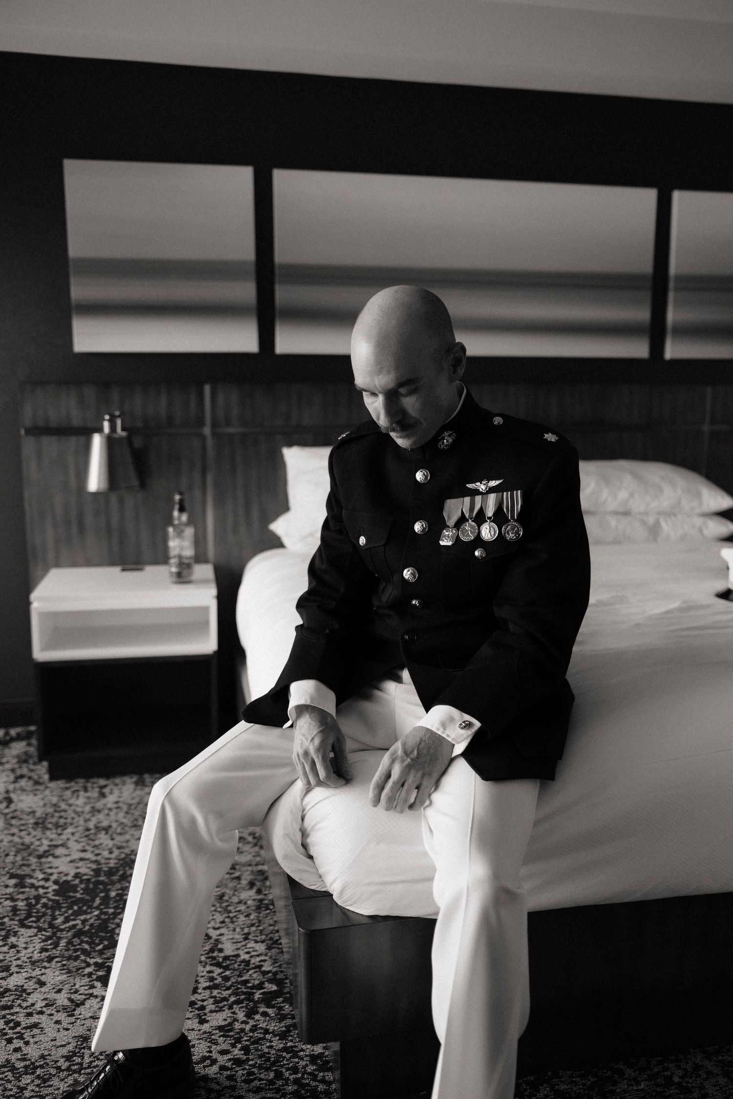 Bald military officer in dress uniform with medals sits pensively on hotel bed in black and white photo.