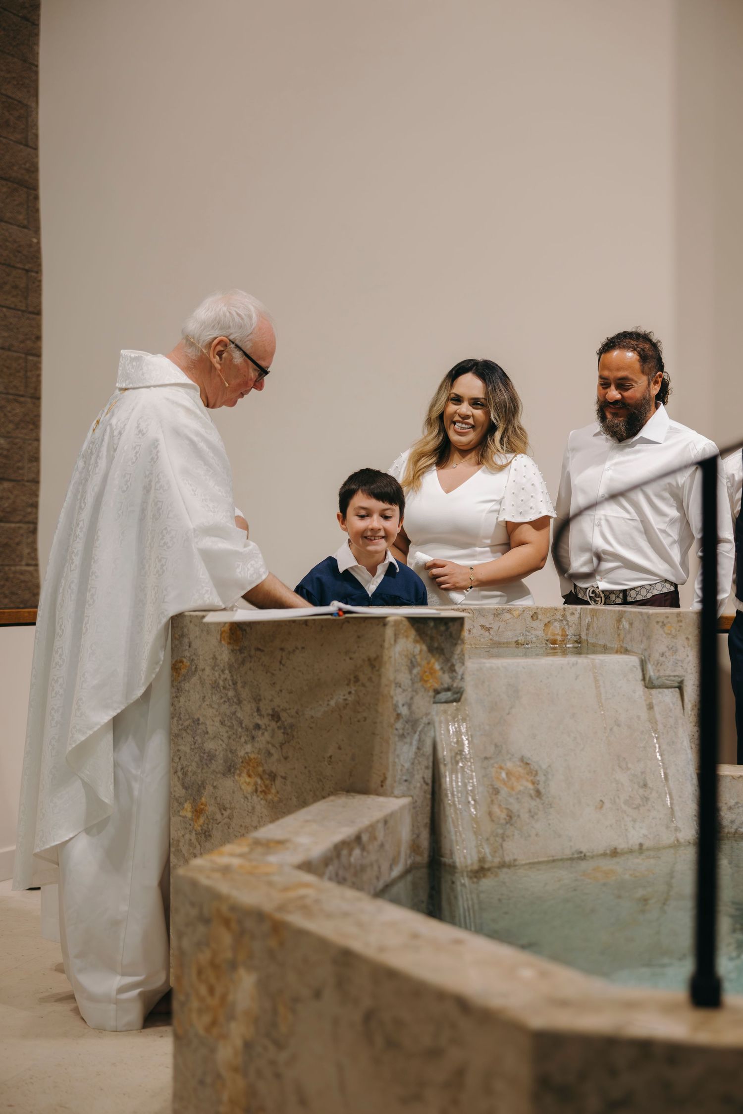 Catholic priest performing a baptism rite with a young boy and his parents at a stone baptismal font.