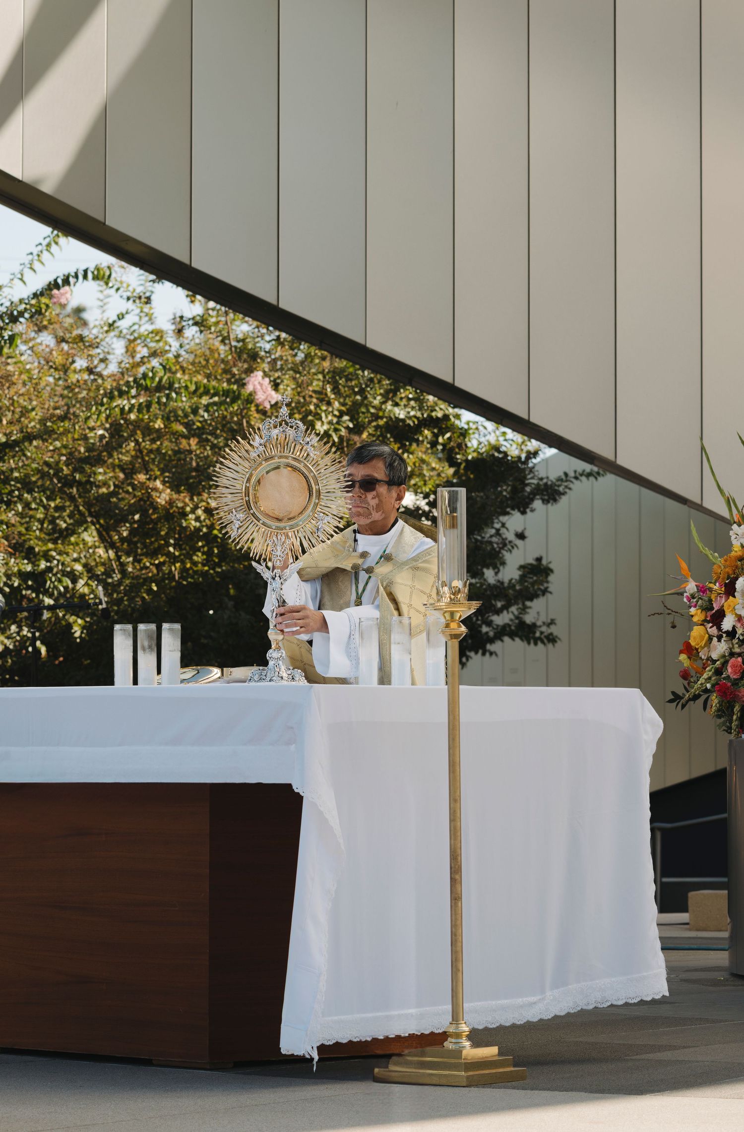 Priest performs benediction with a golden monstrance at a white-clothed altar during an outdoor service.
