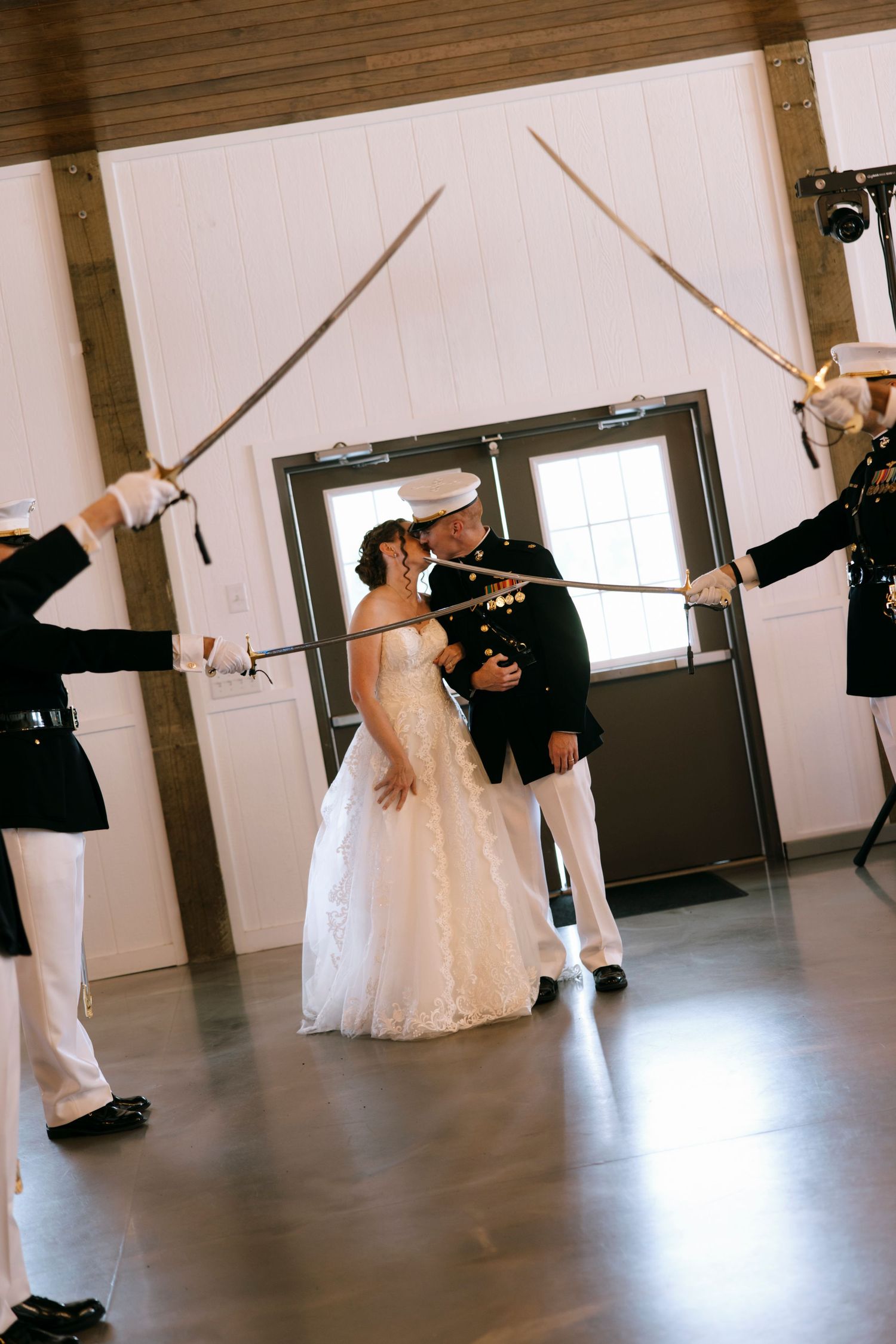 Military wedding couple kissing under arch of swords held by uniformed officers in white dress uniforms inside elegant venue.