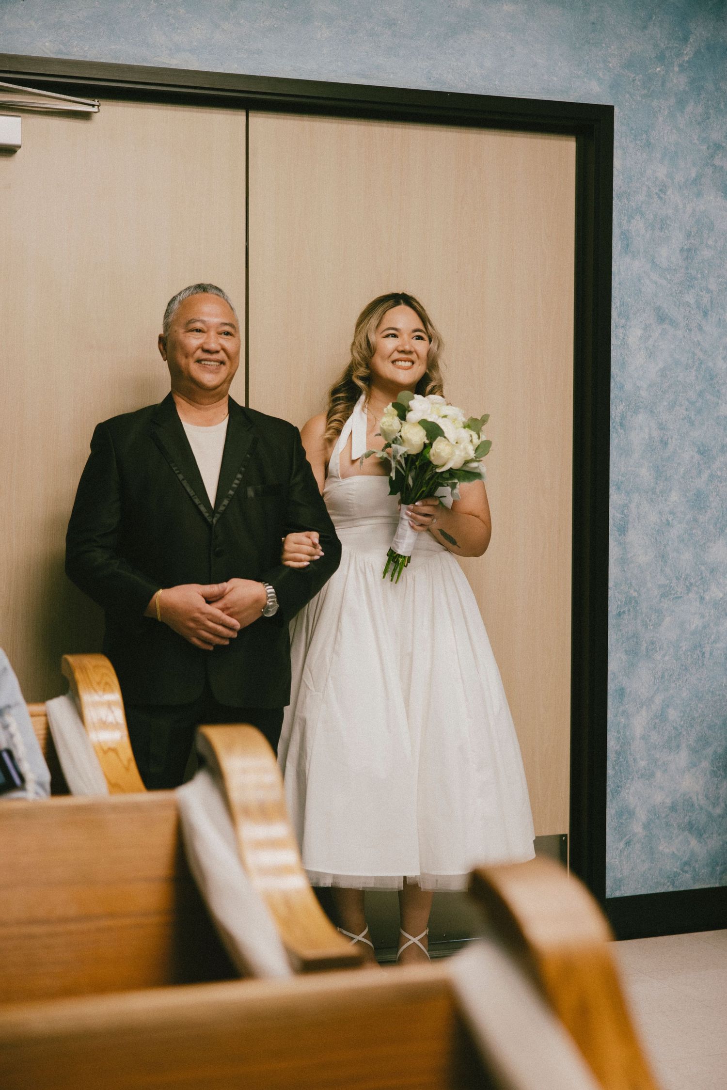 A bride in a tea-length dress holds a white bouquet while standing with someone in formal attire.