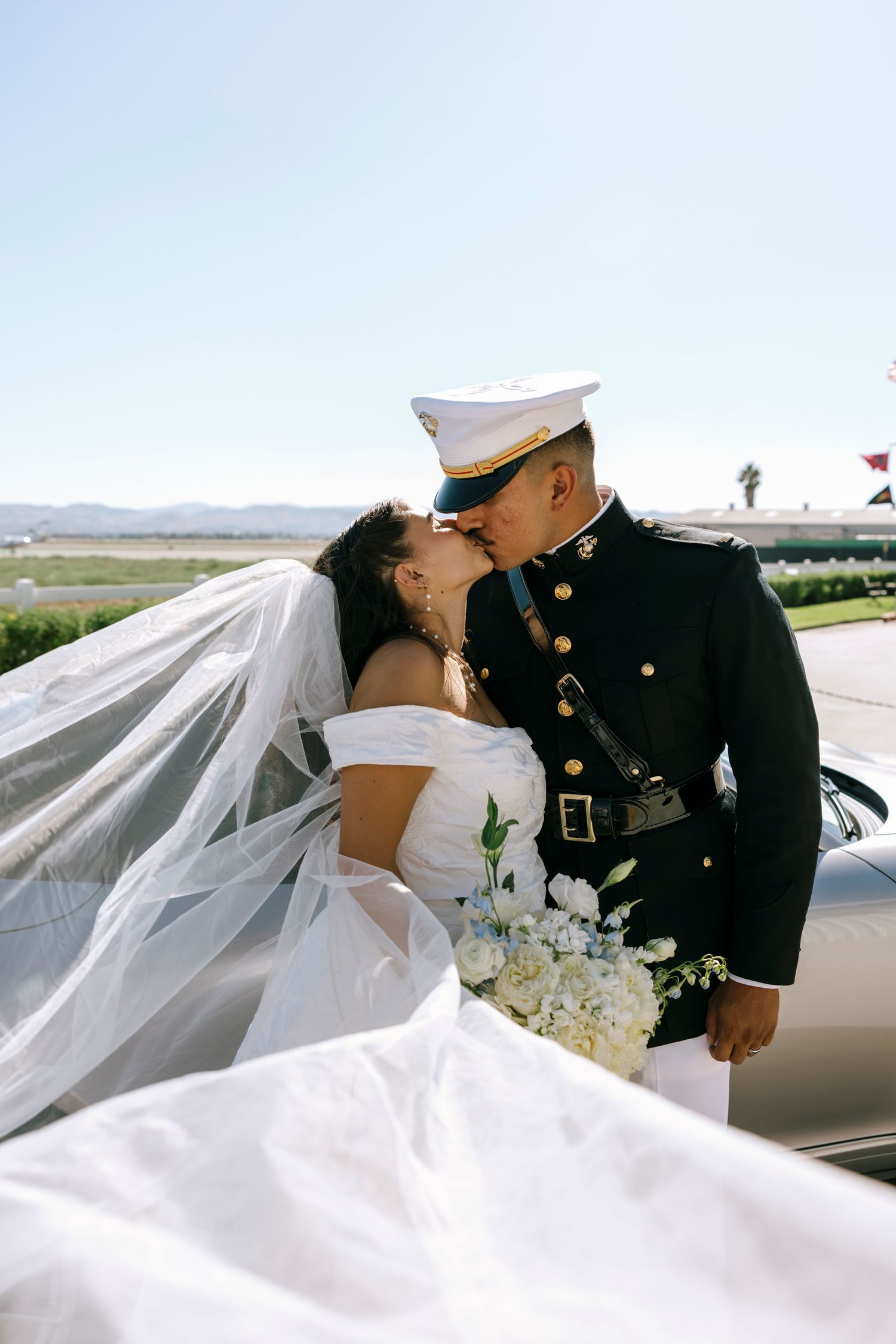 Wedding couple in military dress and white gown embrace near luxury car with veil flowing.