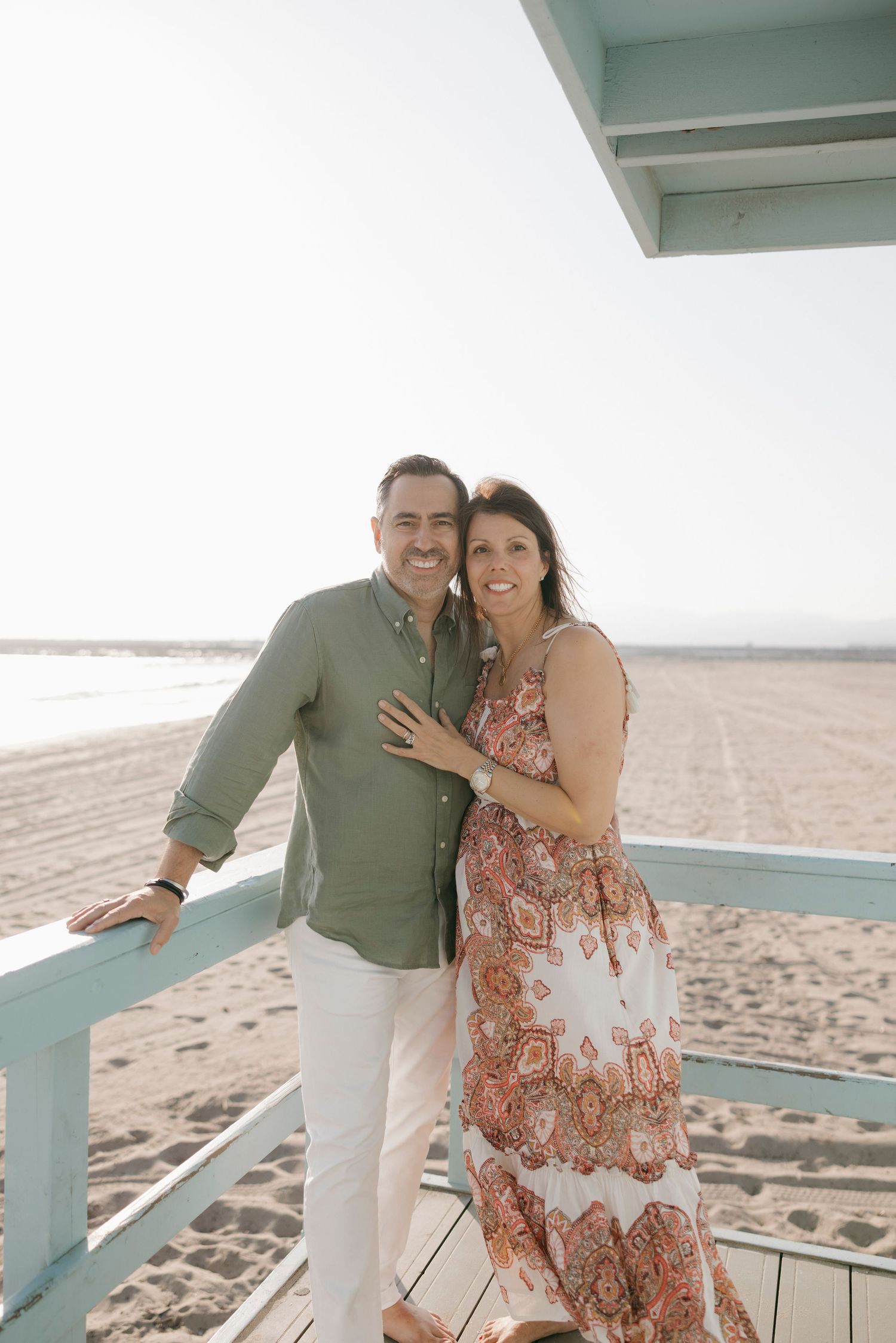 A smiling couple poses on a beach boardwalk, man in green shirt and woman in floral dress, with sandy shore behind them.