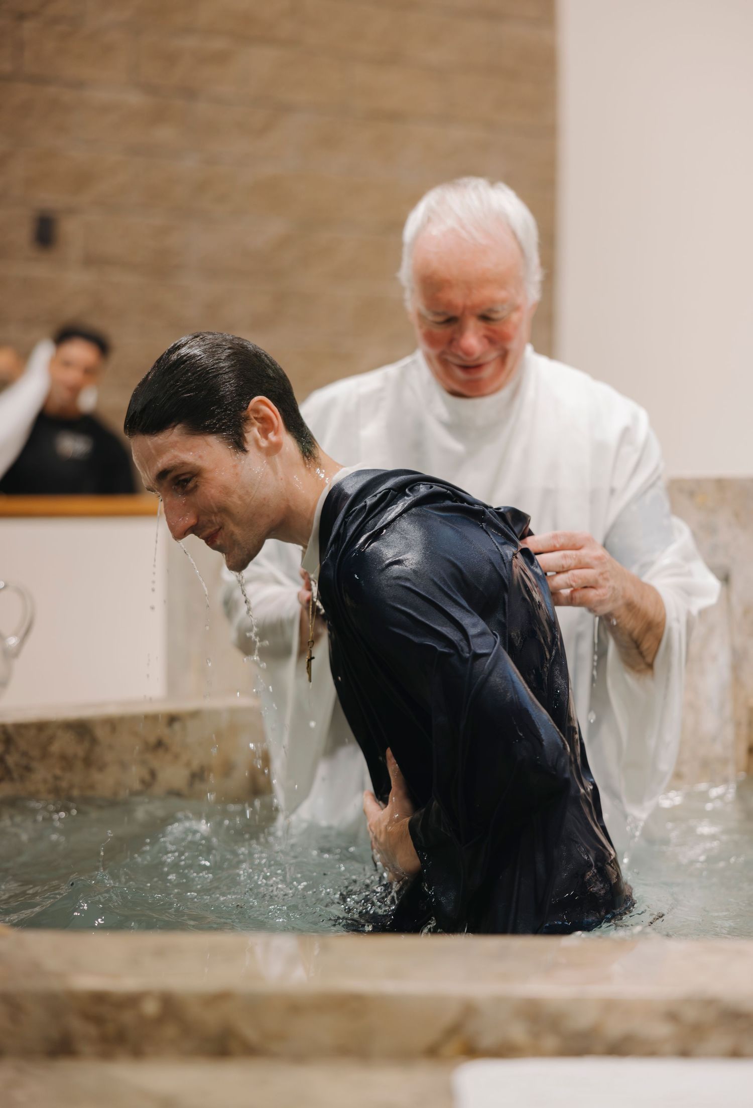 Young man being baptized by full immersion in baptismal pool by elderly priest in white robes.