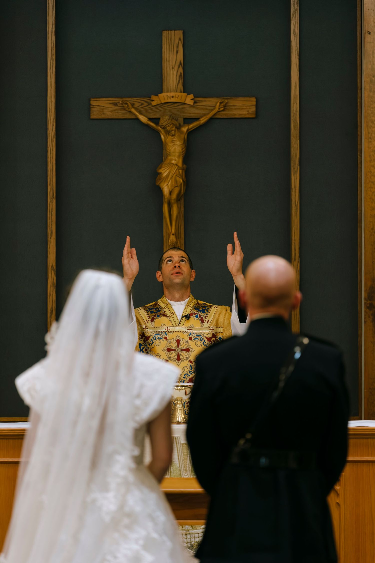 Bride and groom stand before priest with raised hands at Catholic wedding ceremony beneath wooden crucifix.