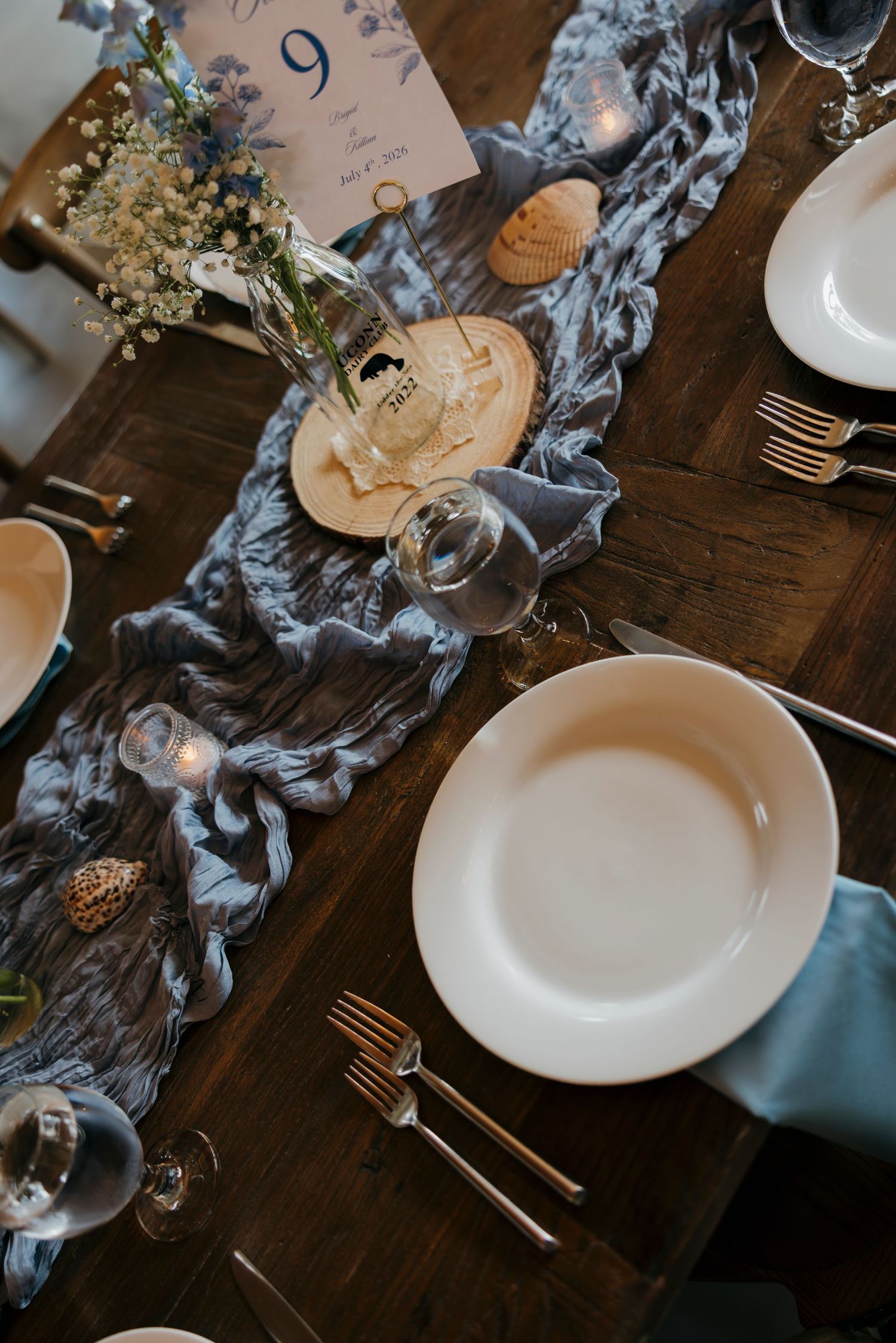 Elegant wedding table setting with blue gauze runner, white plates, gold cutlery, and floral centerpieces on dark wood.