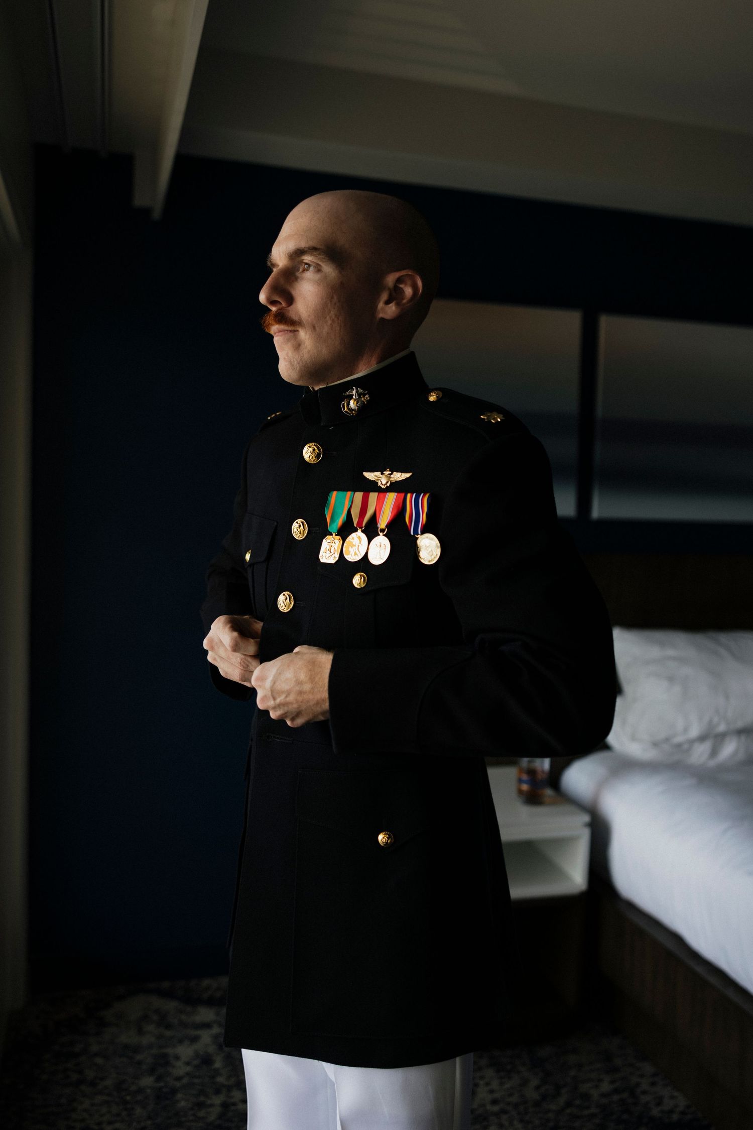 Marine officer in dress blues uniform with medals stands in a hotel room, gazing thoughtfully to the side.