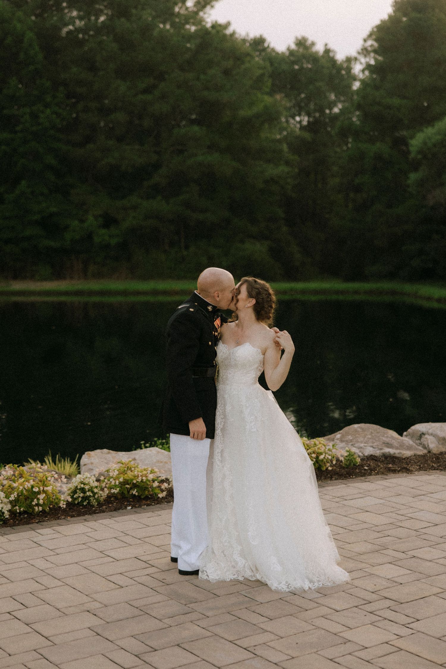 A groom in military dress uniform kisses his bride in a white lace gown by a serene pond surrounded by lush greenery.