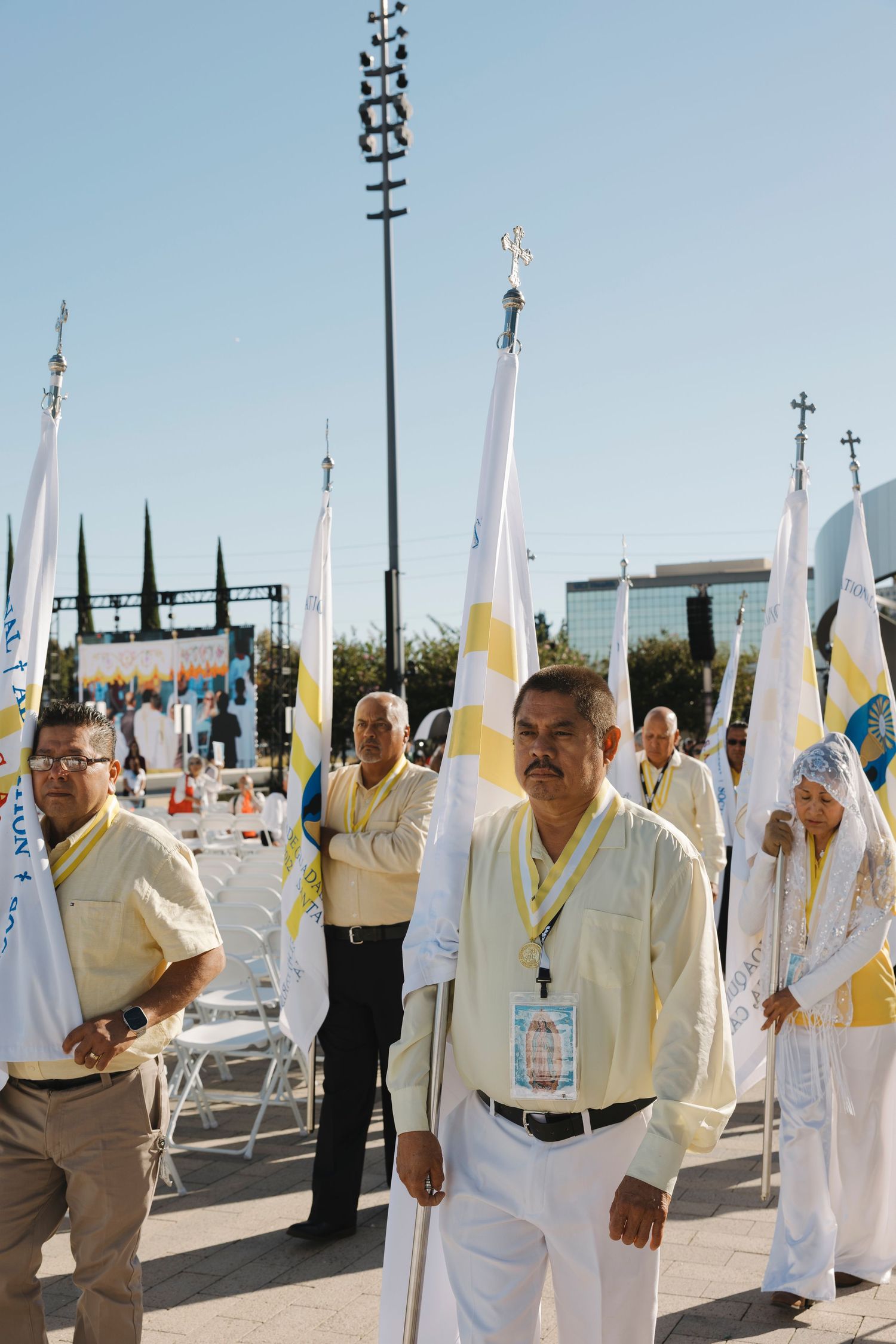 People in white uniforms carrying ceremonial flags at outdoor religious gathering.