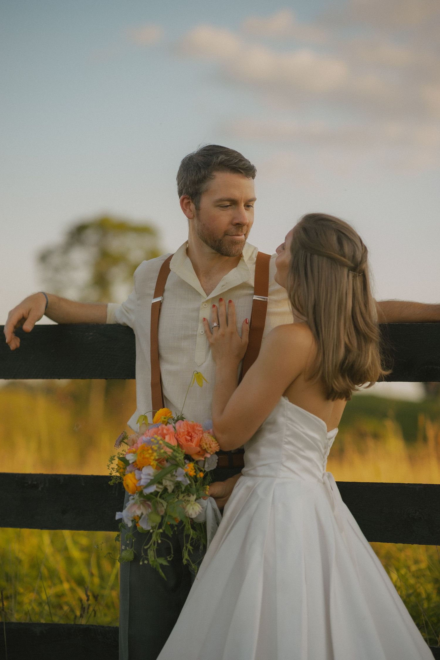 A romantic wedding couple stands by a rustic fence at sunset with a colorful bouquet of flowers.