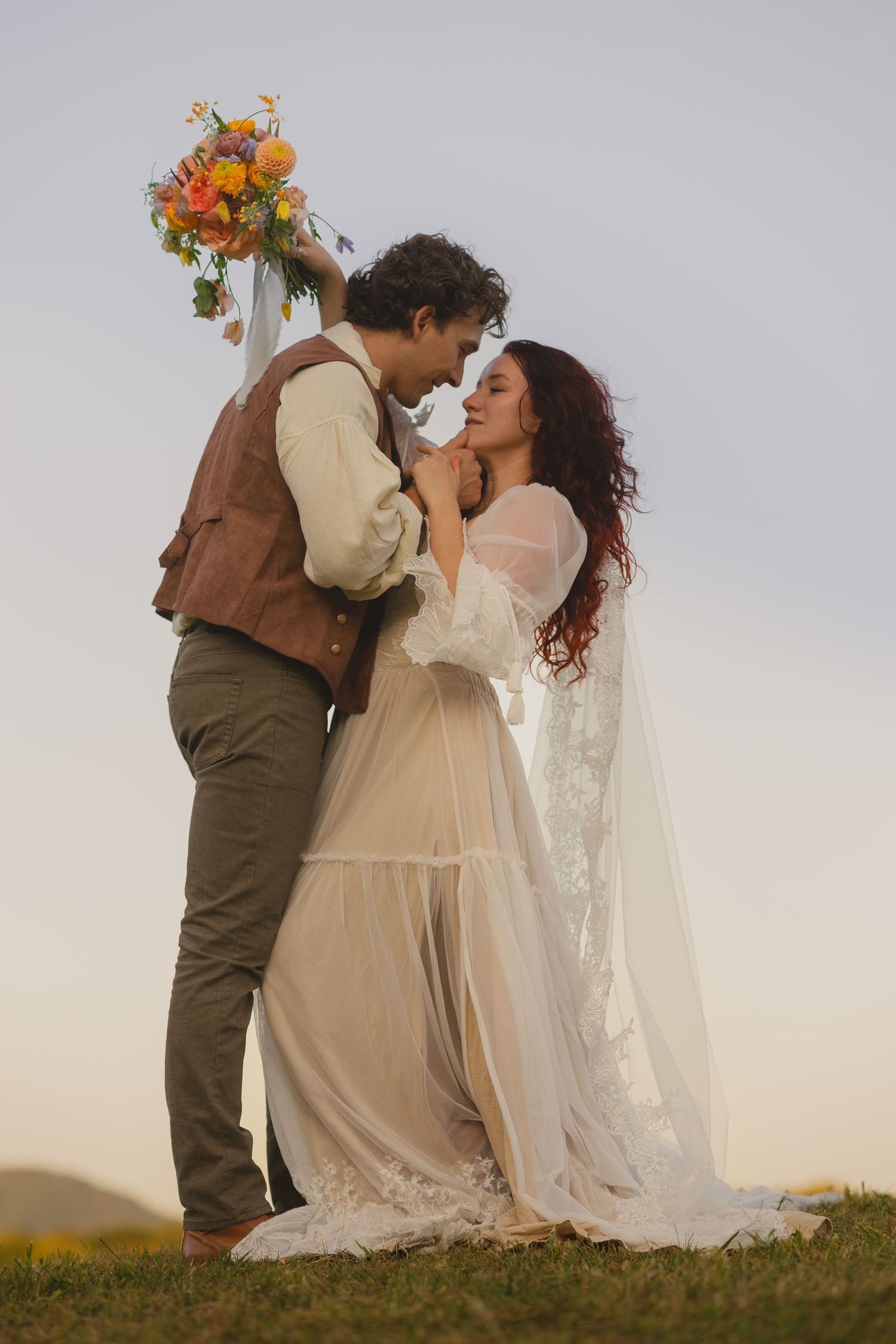 A romantic outdoor wedding scene shows a couple in vintage-inspired attire embracing on a grassy hillside at sunset.