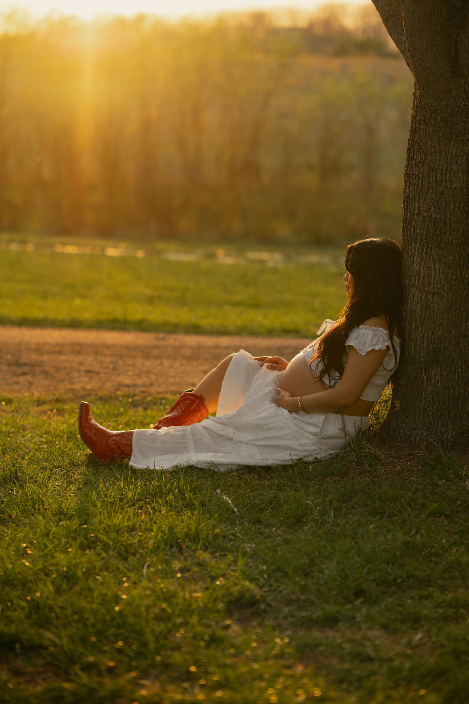 A woman in a white dress sits under a tree at sunset, enjoying the golden light in a peaceful outdoor setting.