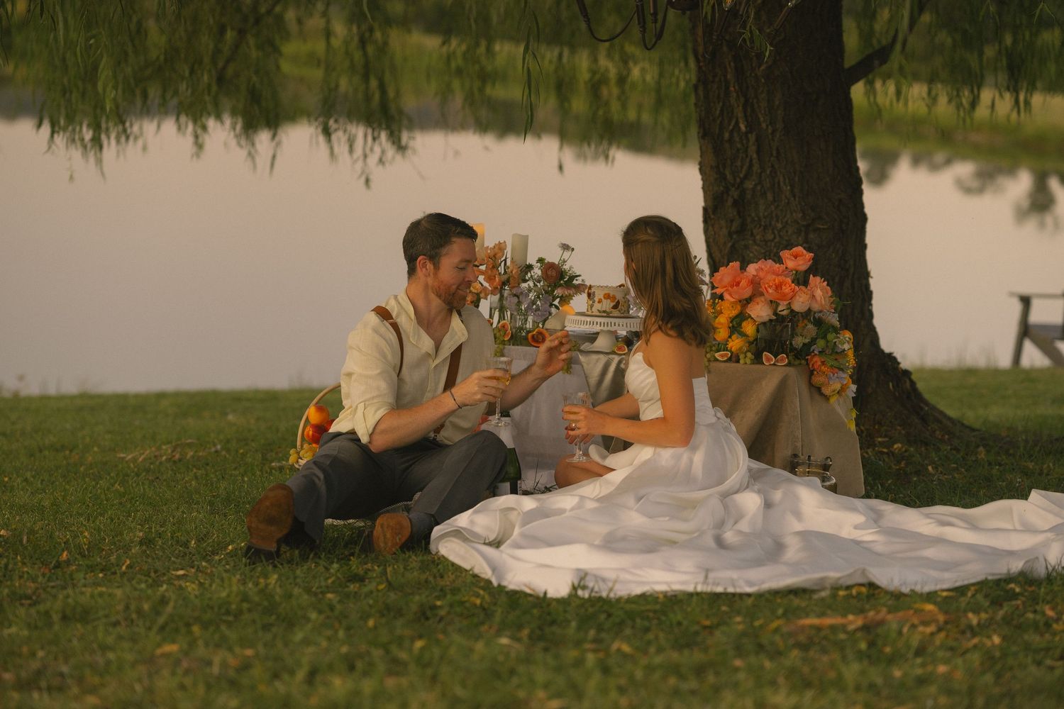 People enjoy a romantic sunset picnic on a grassy lawn with blankets and food spread out beneath a tree.