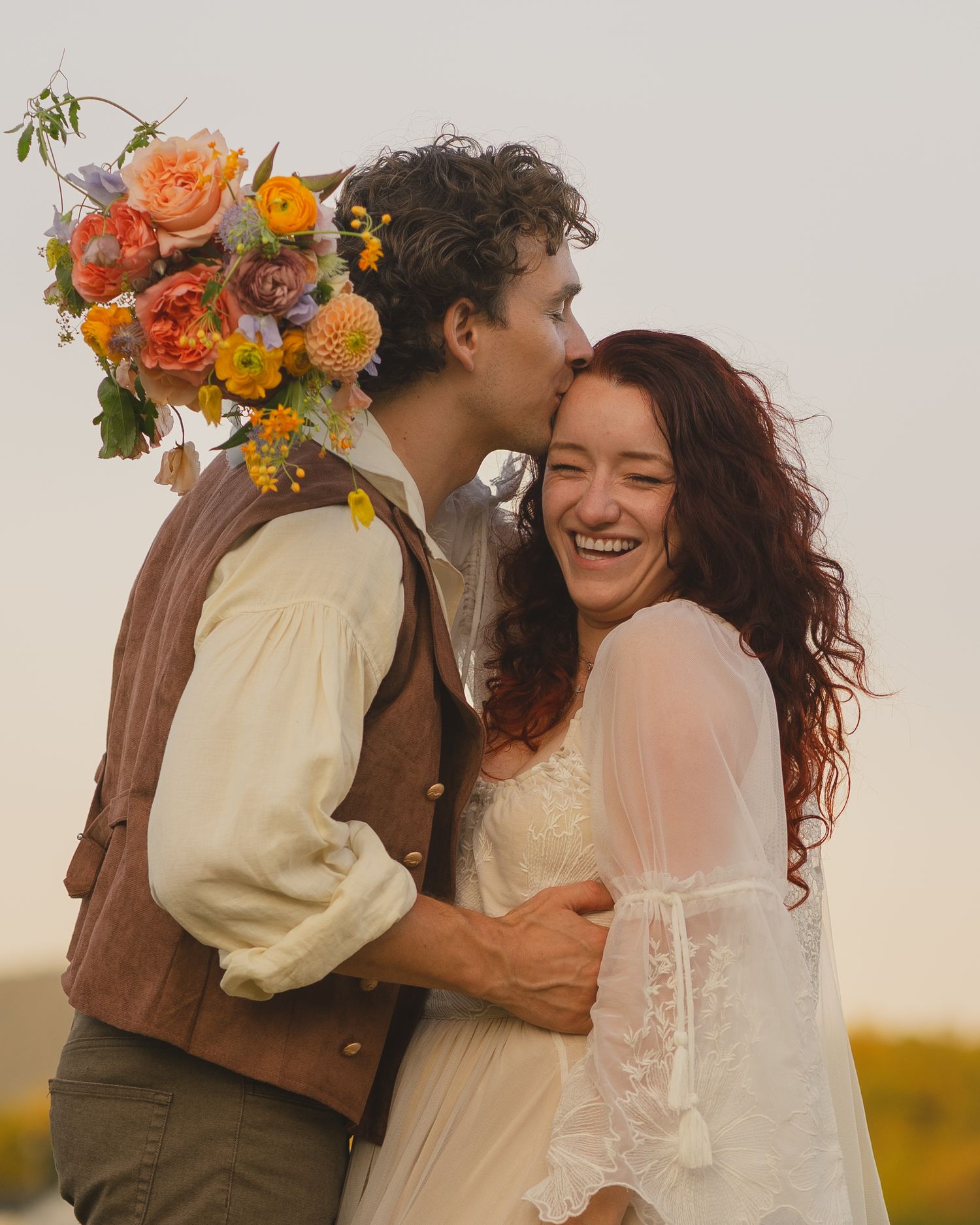 A romantic outdoor wedding scene with a couple embracing surrounded by vibrant orange and yellow floral arrangements at sunset.