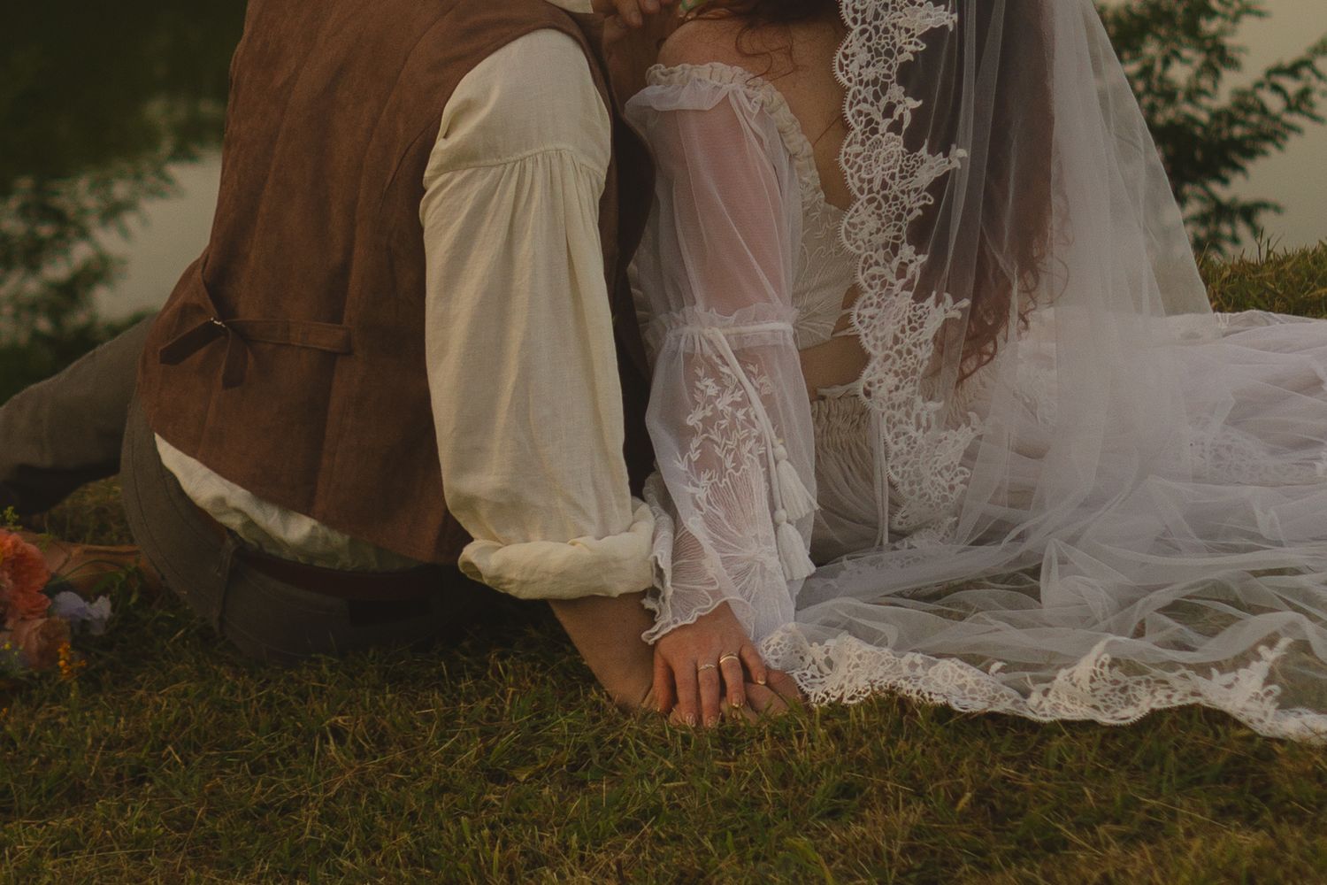 A close-up shot of a bride and groom's hands touching while sitting on grass, with her lace veil flowing beside them.