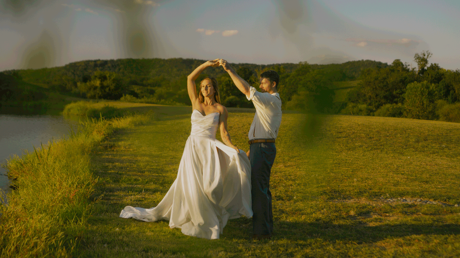 A bride and groom dance joyfully outdoors near a river, surrounded by lush greenery on their wedding day.