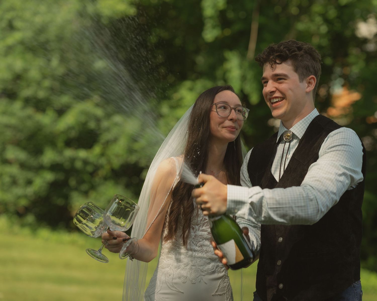 Joyful newlyweds spraying champagne during outdoor wedding celebration.