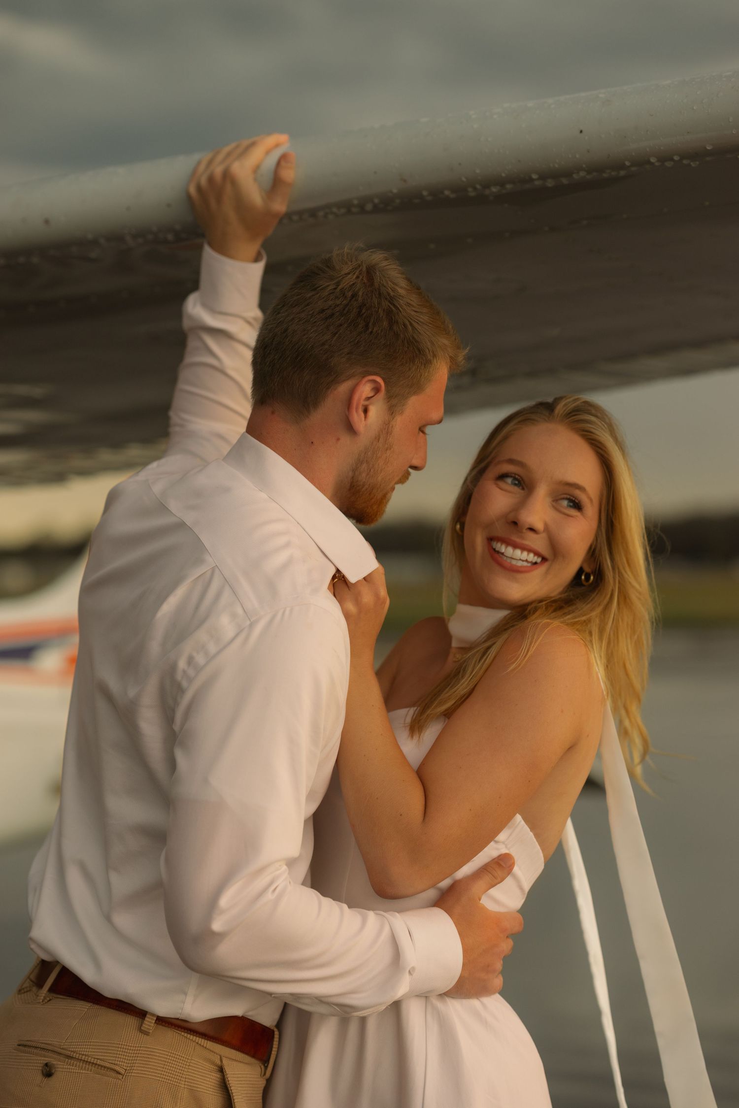 A smiling couple in white outfits embrace near a boat at sunset, sharing a romantic moment by the water.