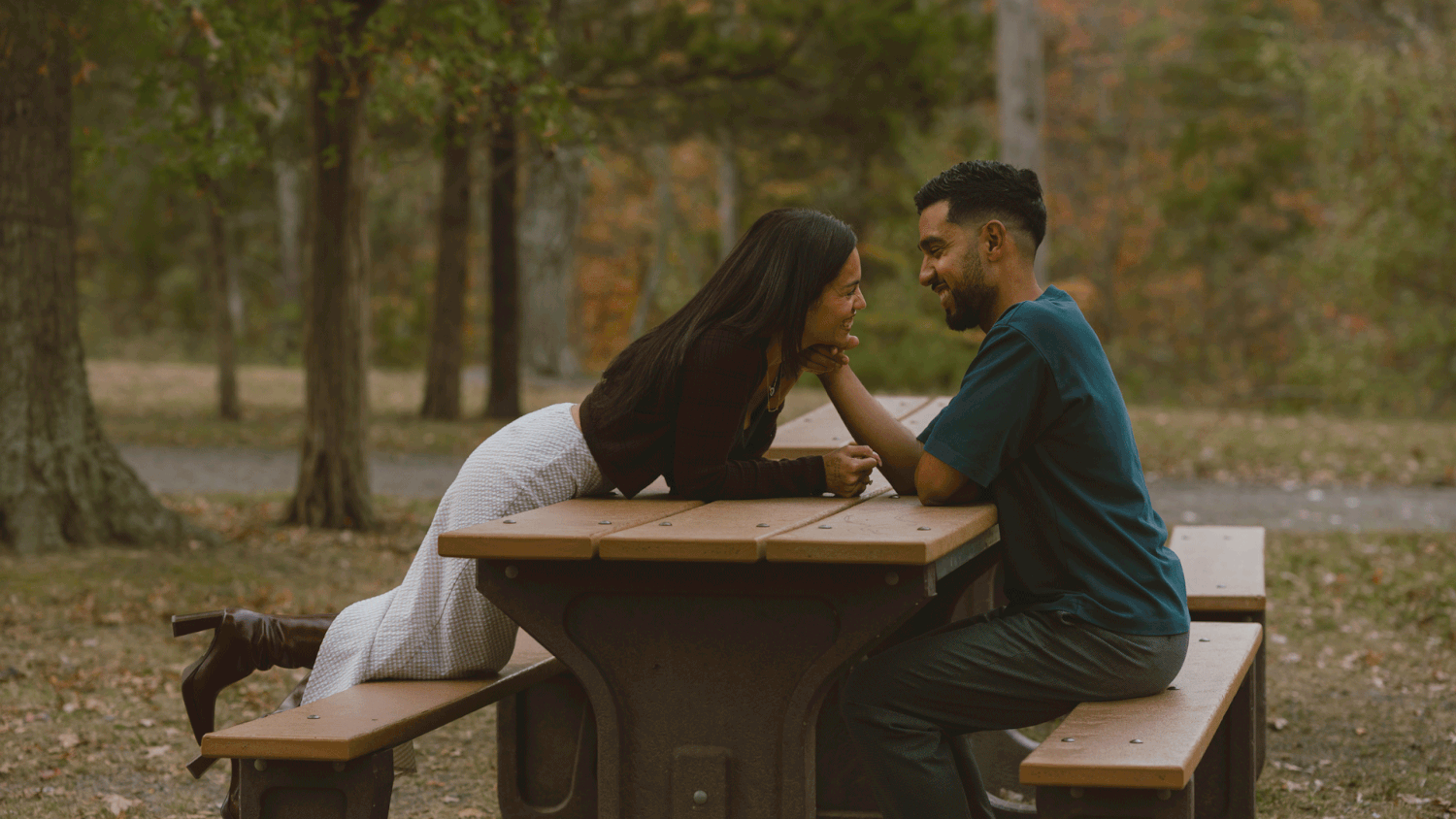 Two people sit together at a wooden picnic table in an autumn park setting.