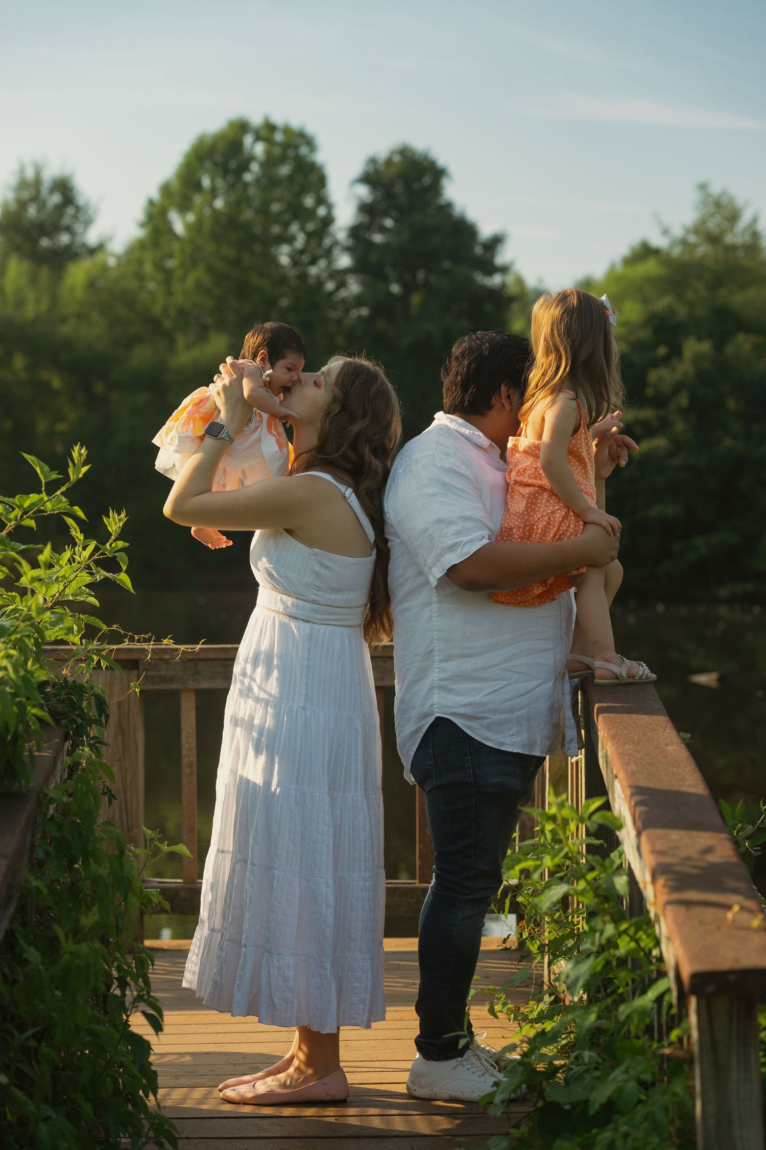 A family shares an intimate sunset moment together on a wooden bridge surrounded by greenery and golden light.
