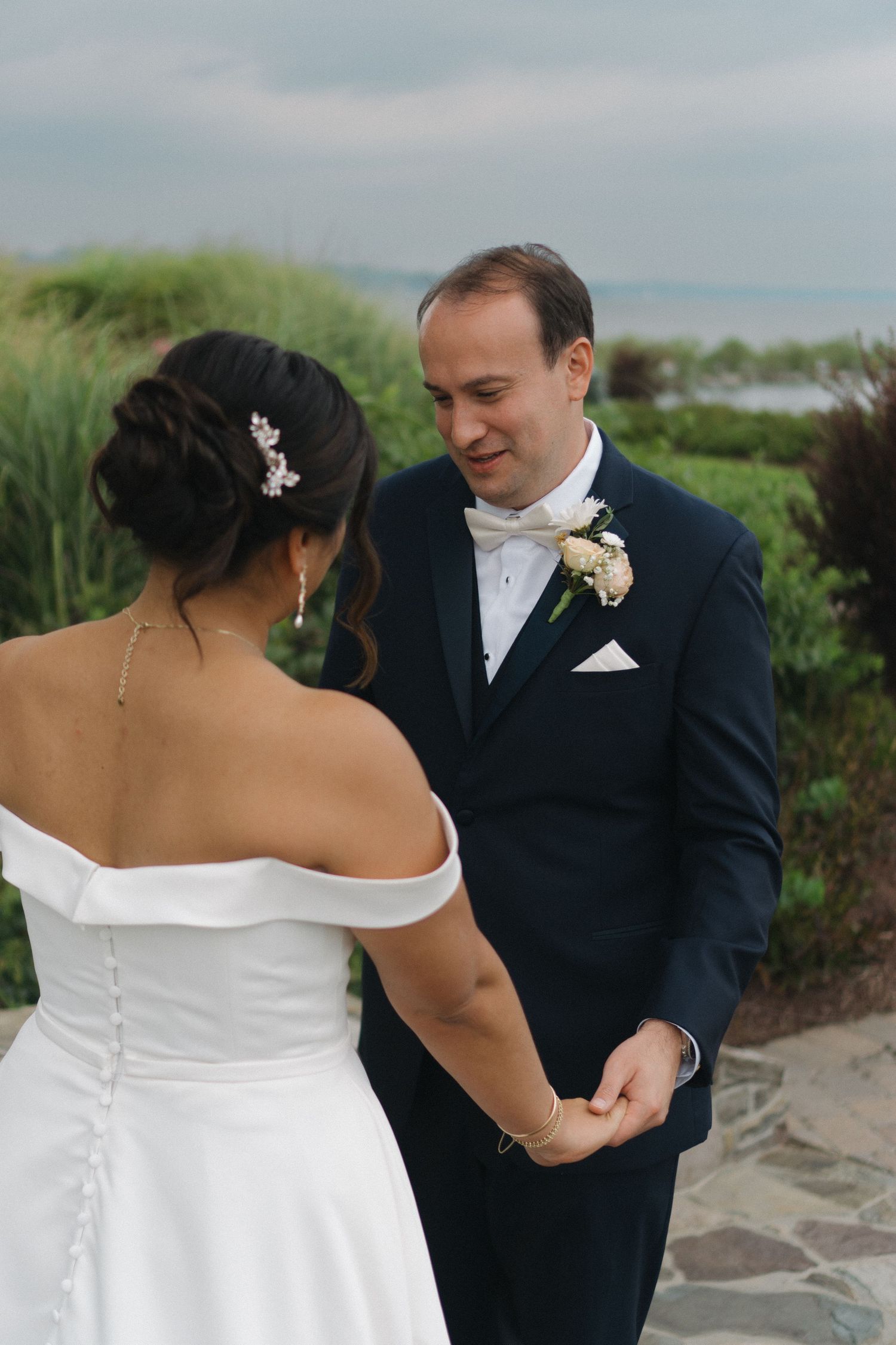 A couple in formal wedding attire share a tender moment together on a beach with greenery in the background.