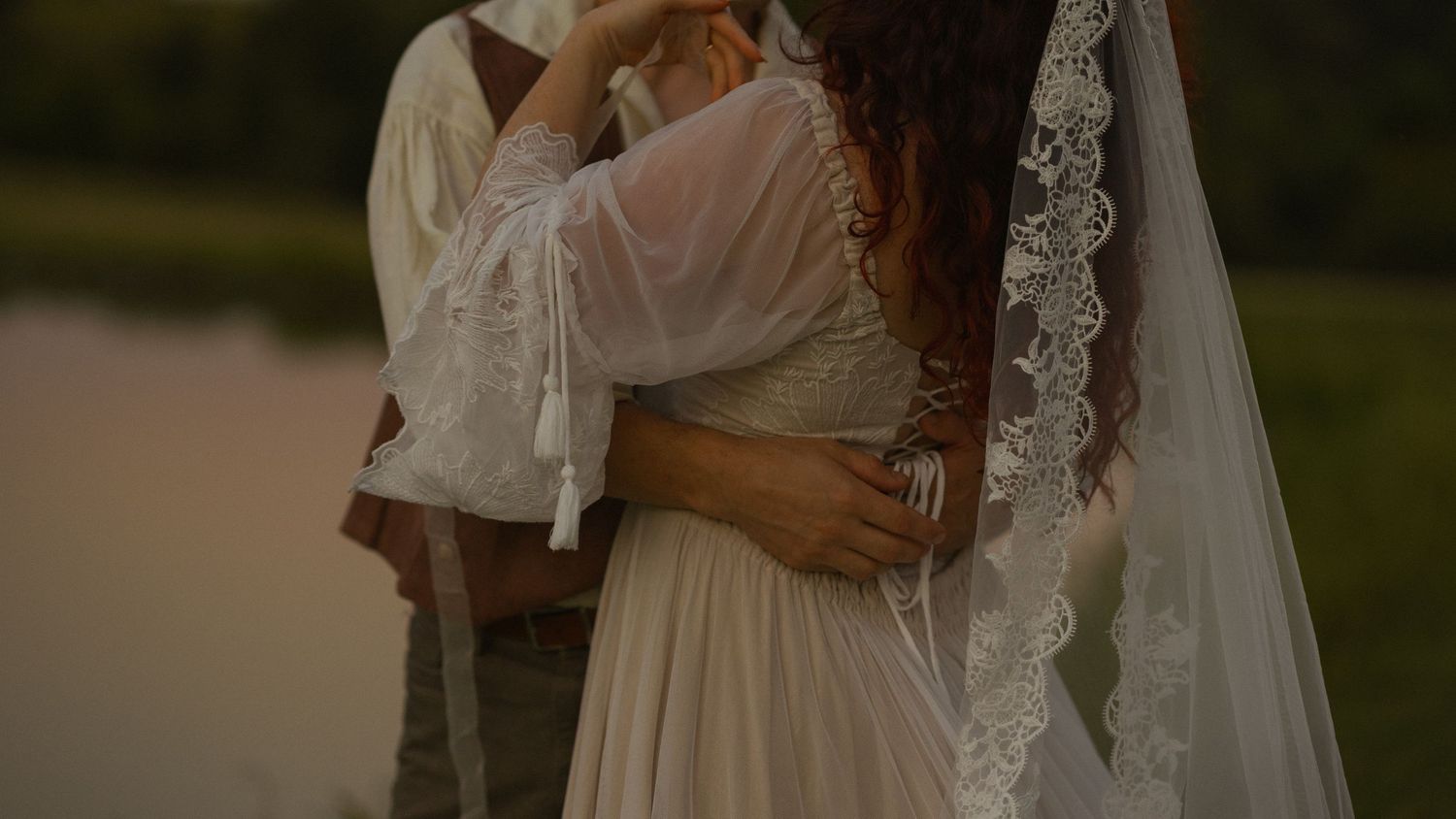 A romantic wedding couple embraces outdoors while wearing a white dress and suit at sunset.