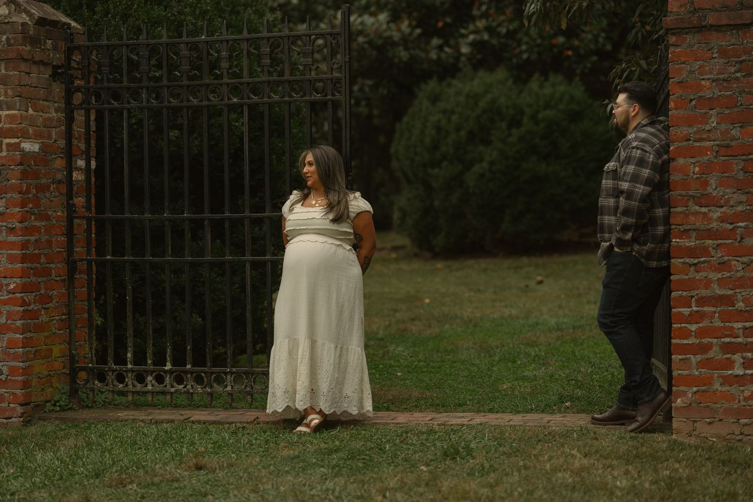 Two people stand near a brick wall entrance with iron gates in an outdoor garden setting.