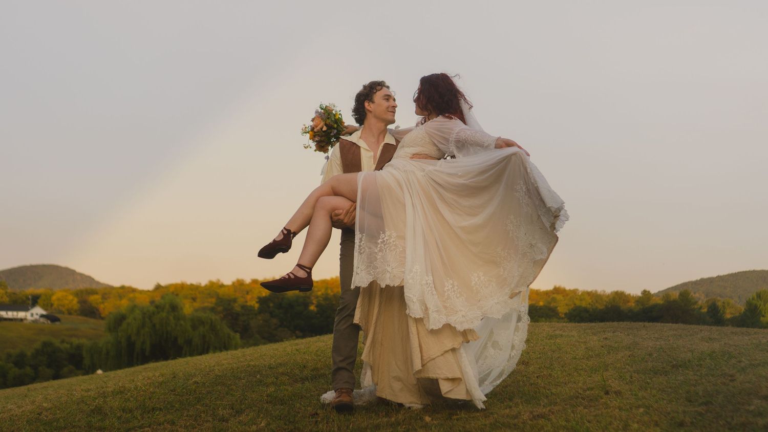 Two brides celebrate outdoors on a green hill with a rainbow and golden trees in the background at their wedding.