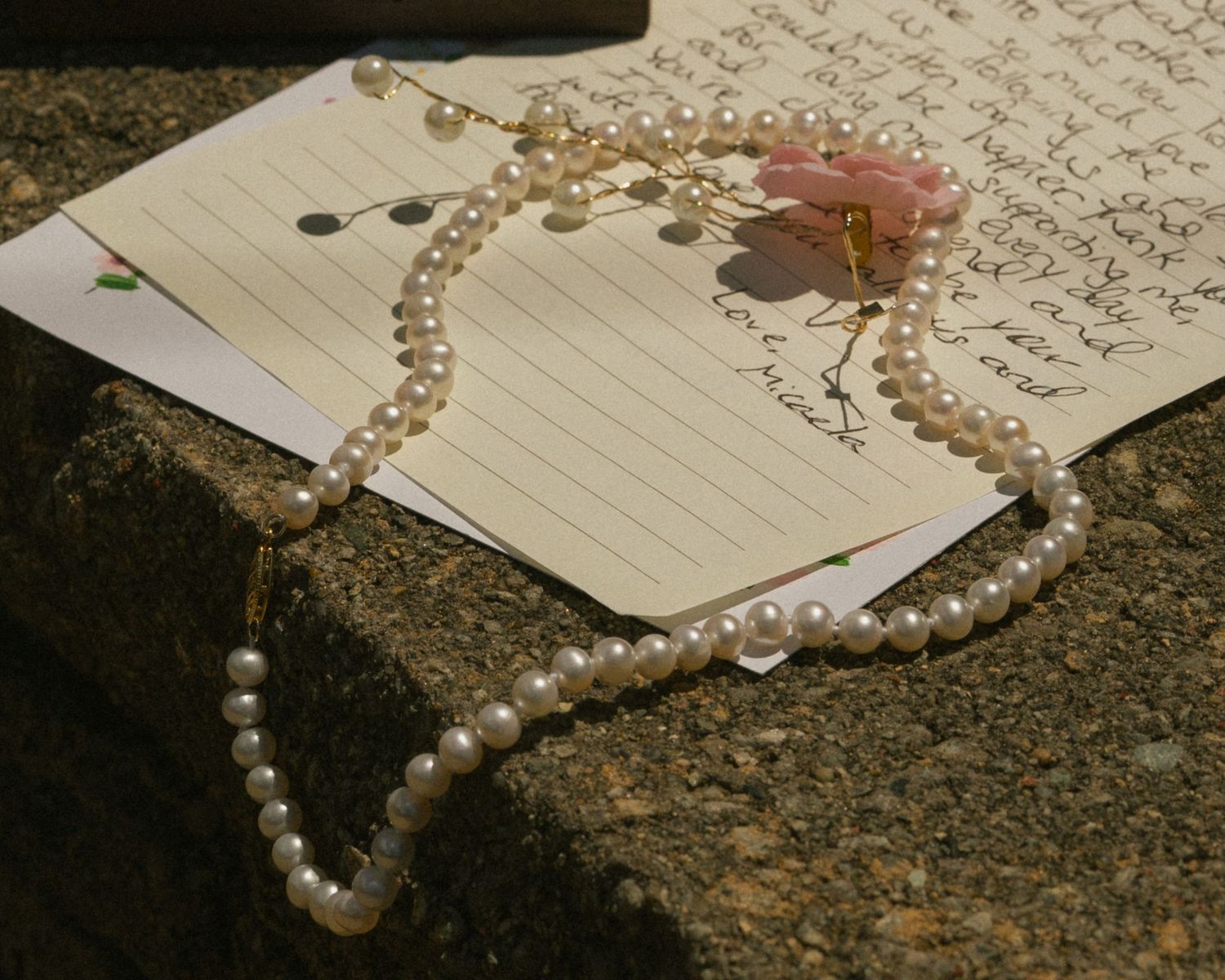 Pearl necklace and beaded hair accessory with pink flower resting on handwritten letters on granite surface.