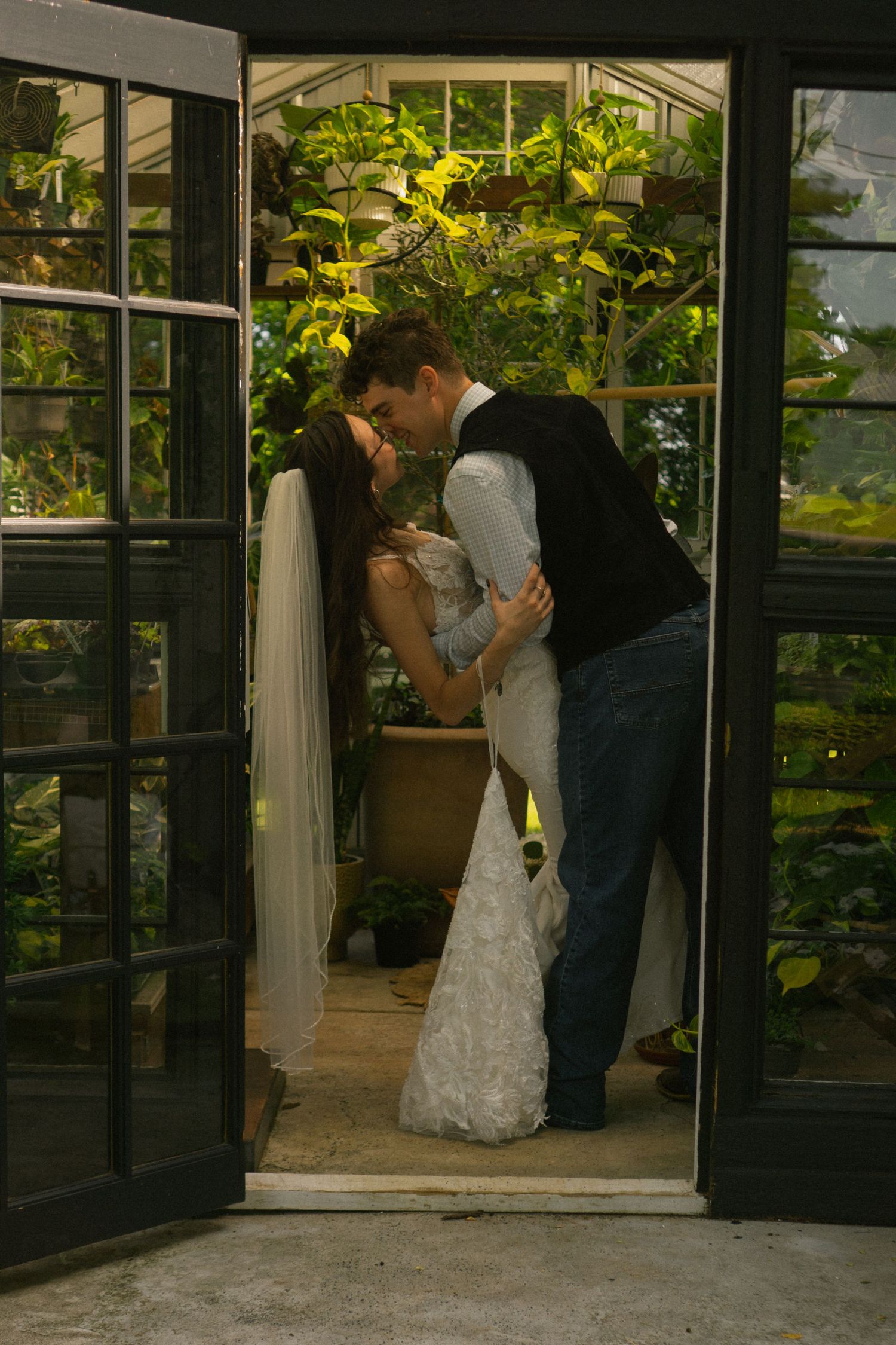 Bride and groom sharing a romantic kiss in a lush greenhouse doorway, bride wearing a white lace gown and veil.