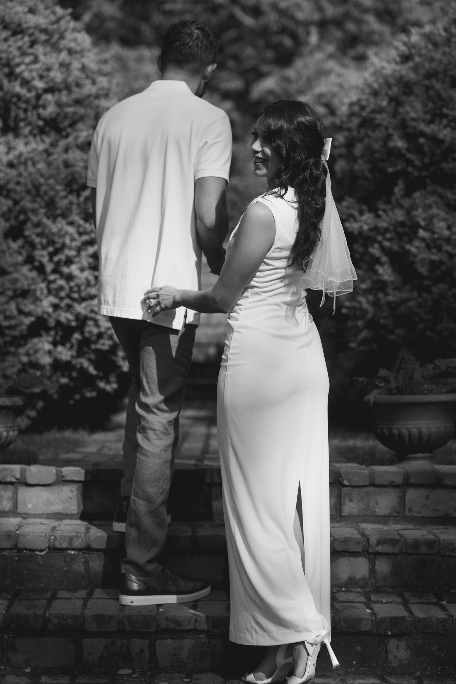 A romantic black and white photo series showing a couple in formal attire embracing by a stone wall in a garden setting.