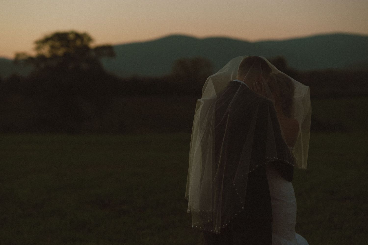 Newlyweds kissing underneath the bride's veil while the sun dips behind the Blueridge Mountains