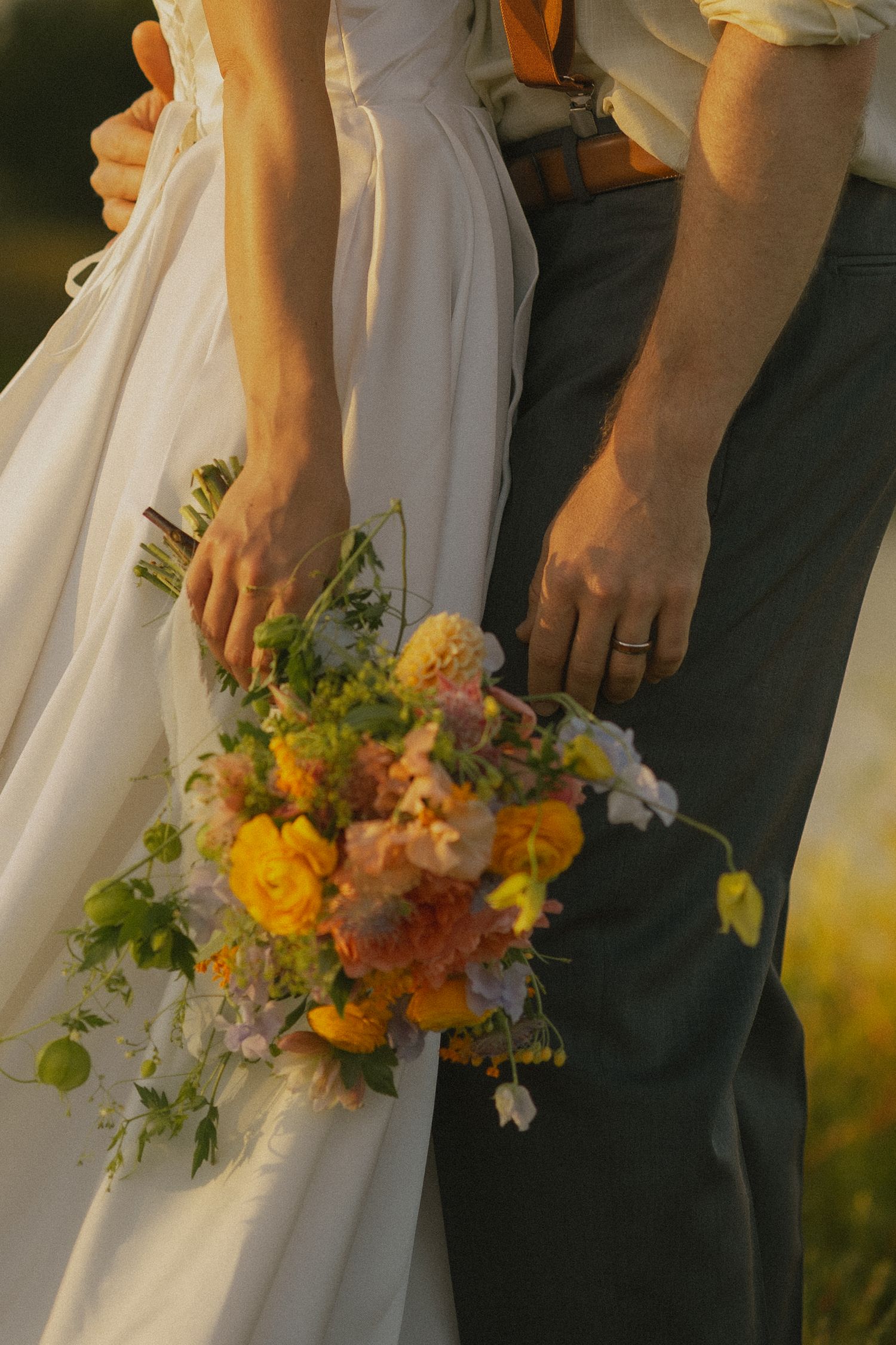 Wedding couple holds colorful summer bouquet with yellow and peach flowers against dark suit and white dress.