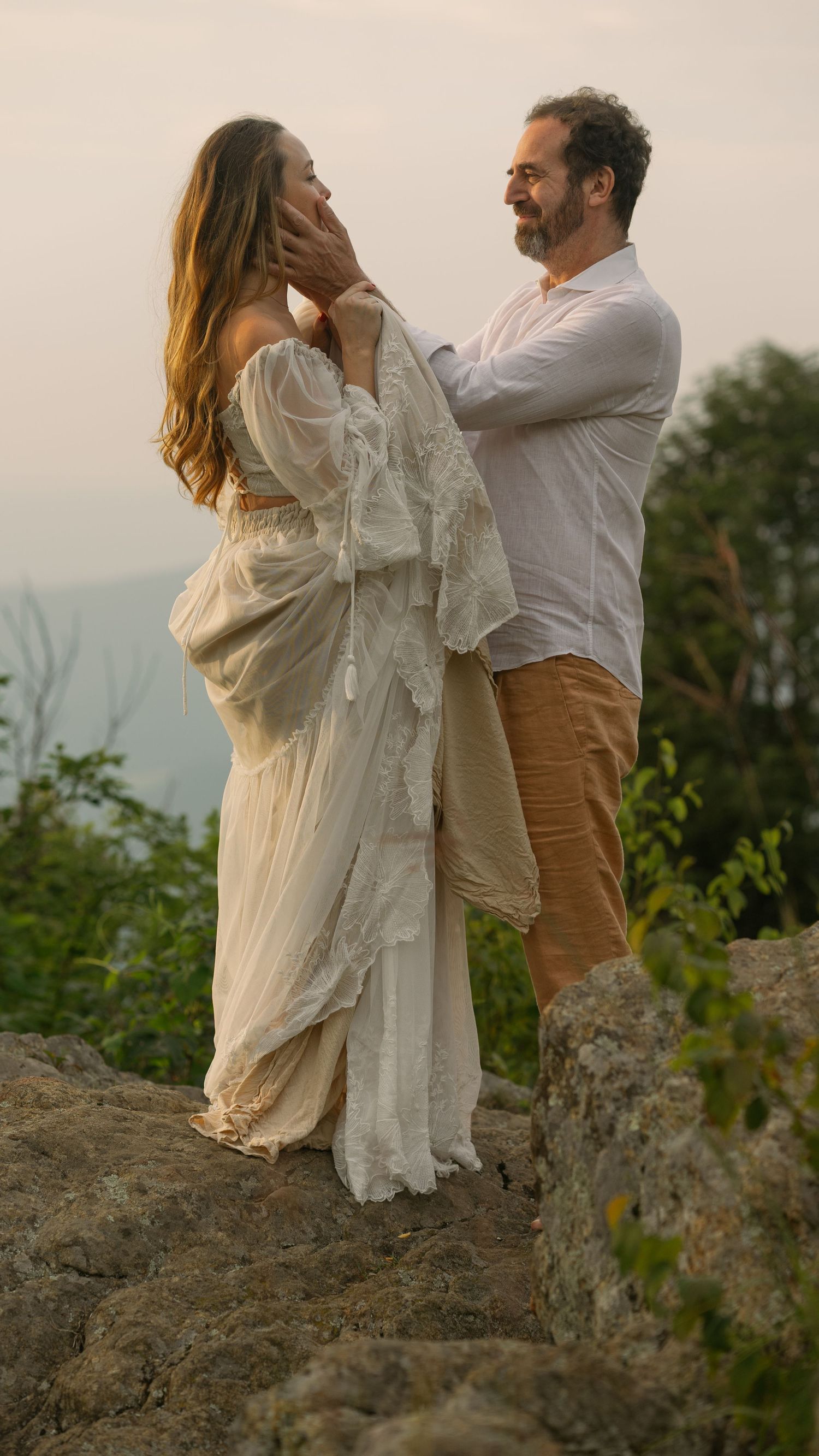 A couple in white clothing shares an intimate moment while standing on a rocky overlook at sunset.