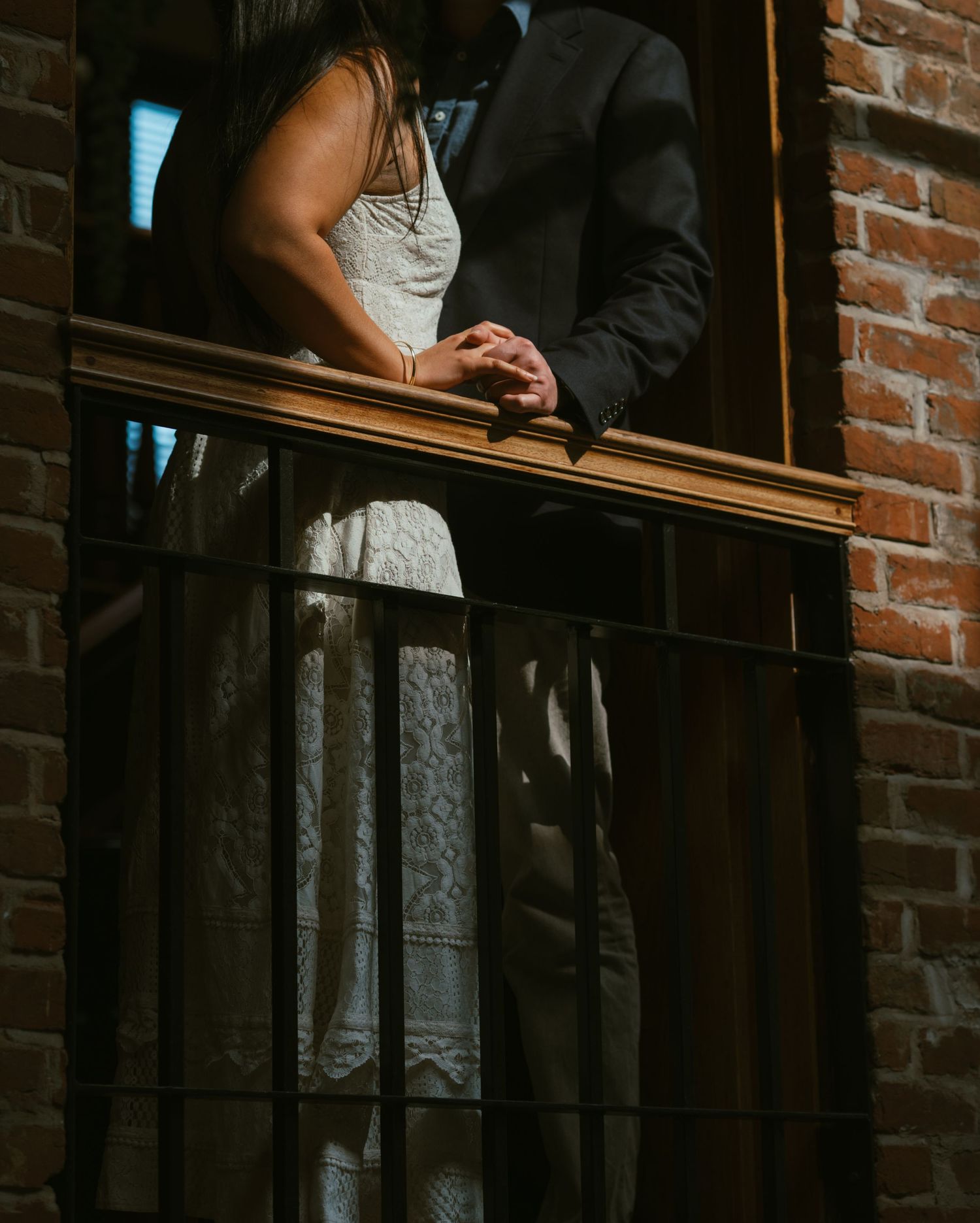 Couple in formal attire standing against exposed brick wall in moody lighting.